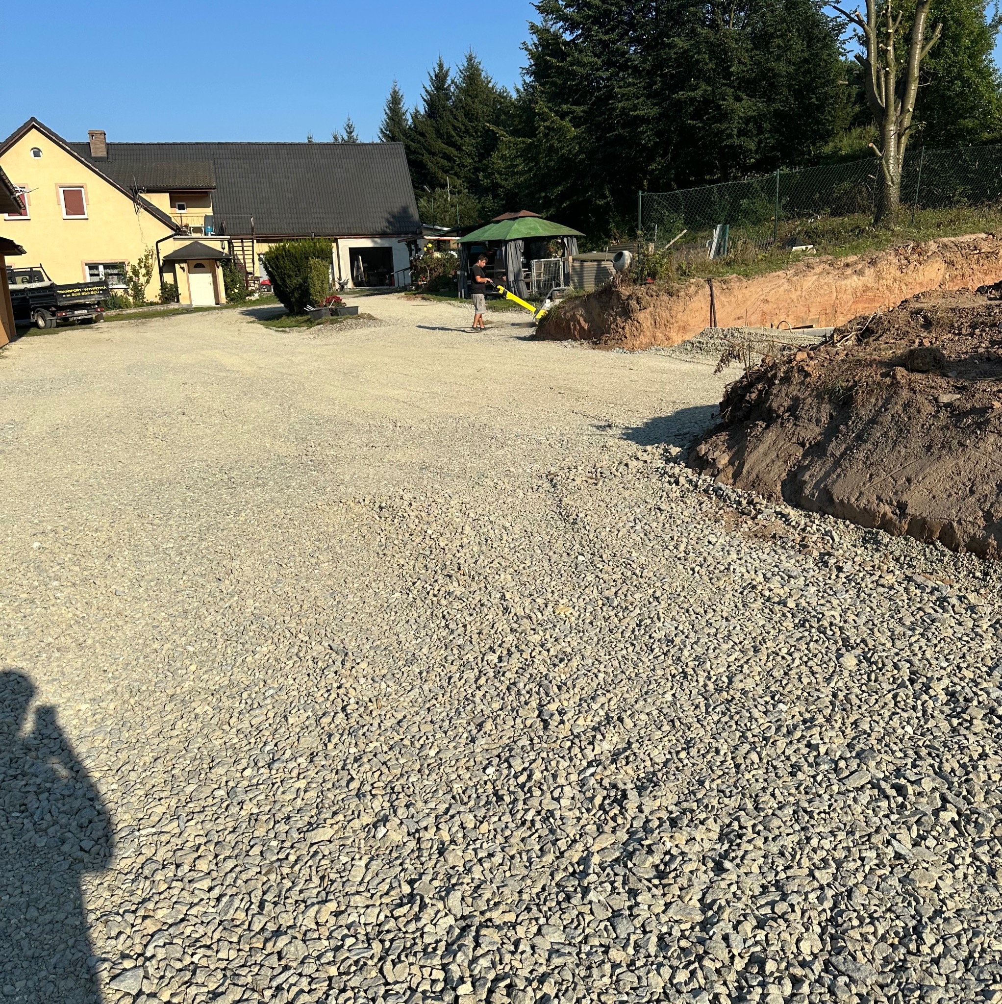 Nierównomiernie rozłożony tłuczeń na placu budowy, widoczny pracownik z zagęszczarką w tle, wykop ziemny z boku.