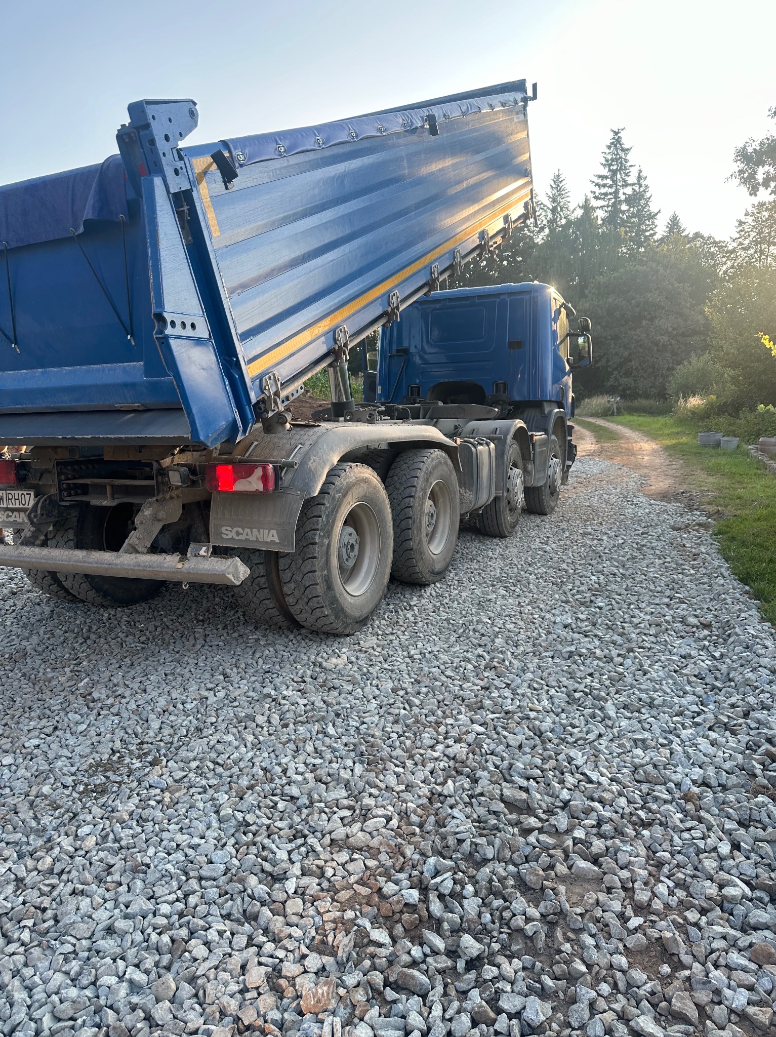 Ciężarówka Scania z podniesioną skrzynią ładunkową wysypuje szarą frakcję kamienną na gruntową drogę, widoczne drzewa w tle.