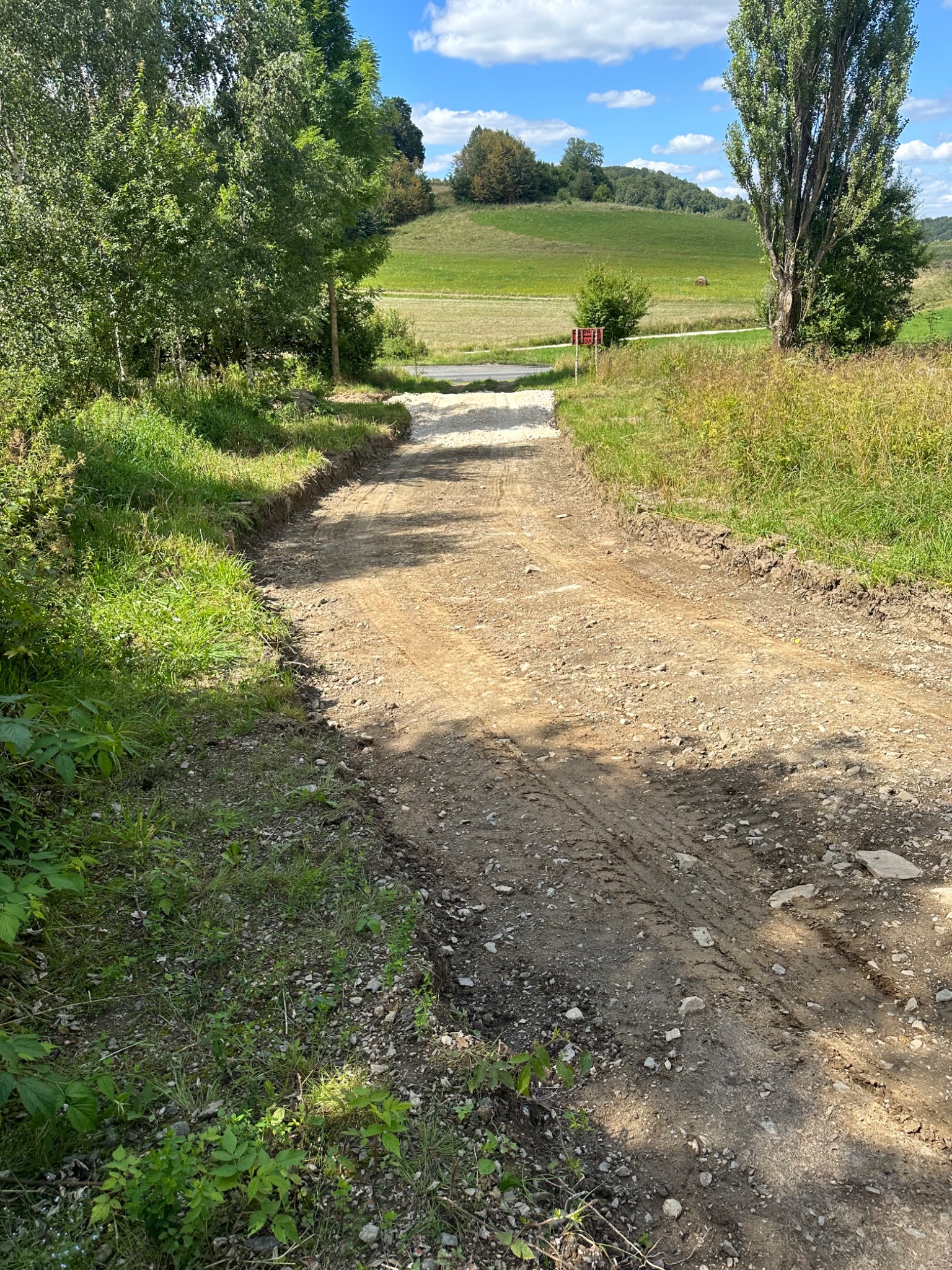 Droga gruntowa w trakcie prac, widoczne ślady opon, wykarczowane pobocza, w tle zielone pola i drzewa.