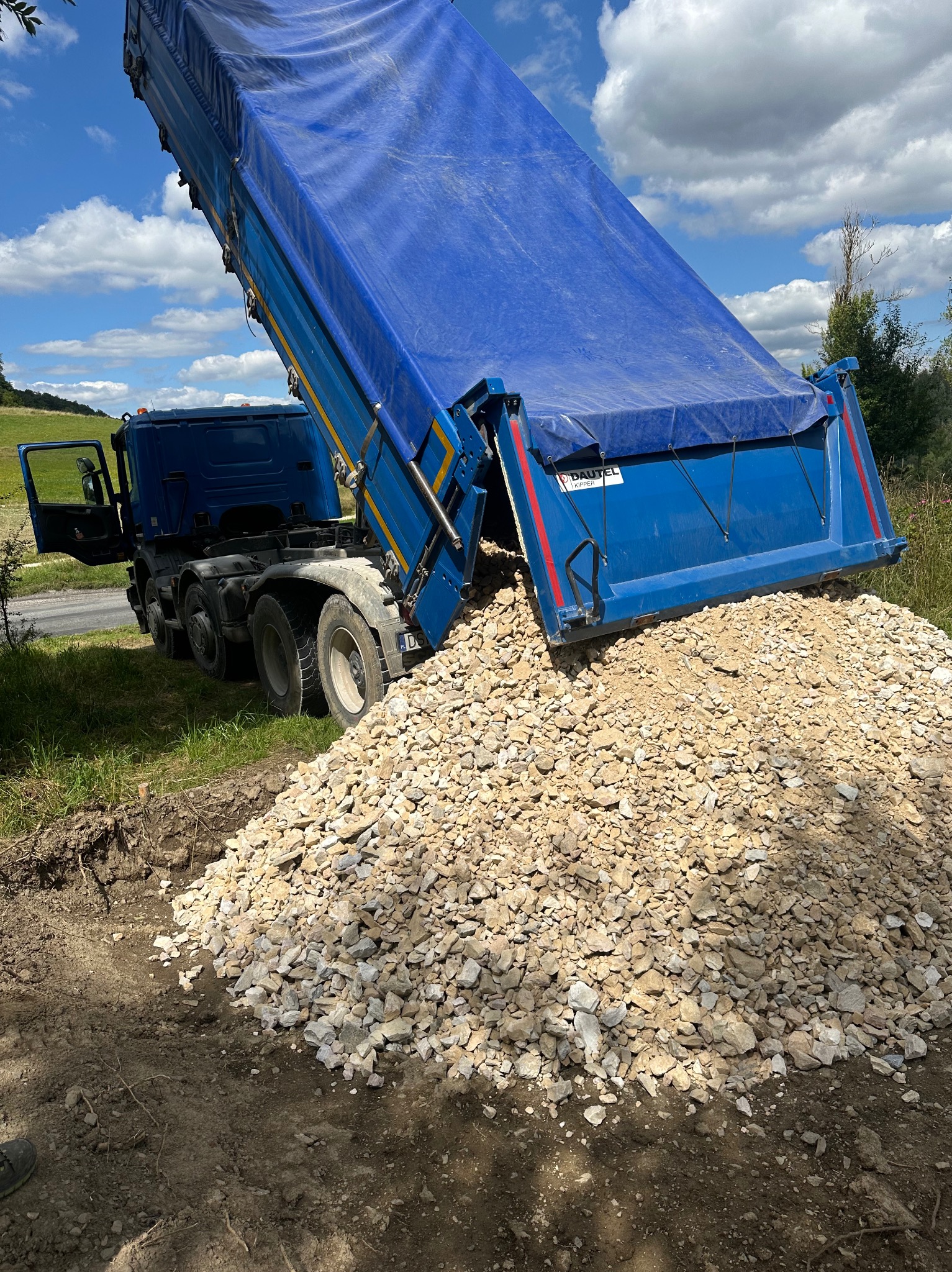 Ciężarówka z niebieską plandeką wysypuje jasny gruz na ziemię, widoczny fragment buta w lewym dolnym rogu kadru.