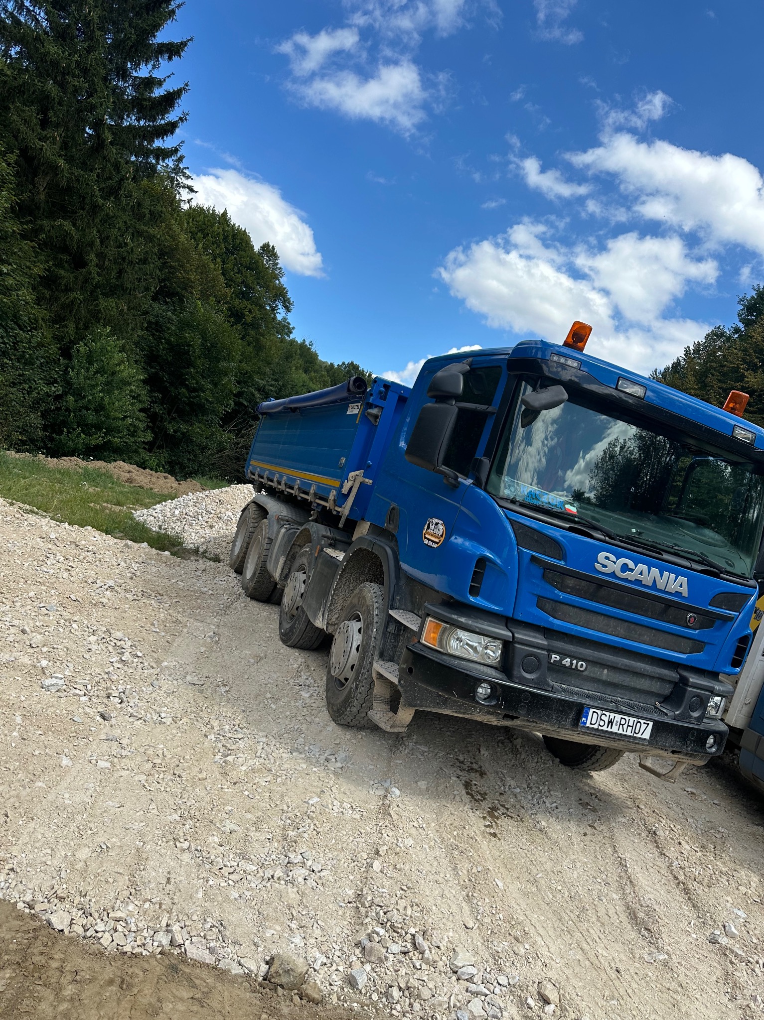 Niebieska wywrotka Scania P410 na drodze z kruszywa, las w tle i błękitne niebo z chmurami.
