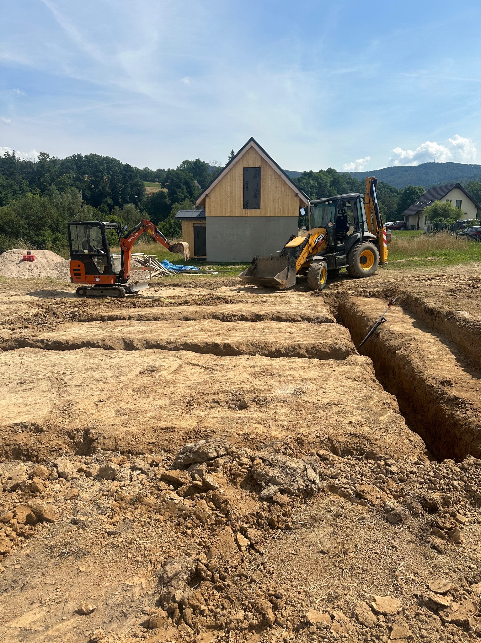 Dwa traktory kopiące rowy w ziemi pod budowę domu w słoneczny dzień, w tle las i inne domy.