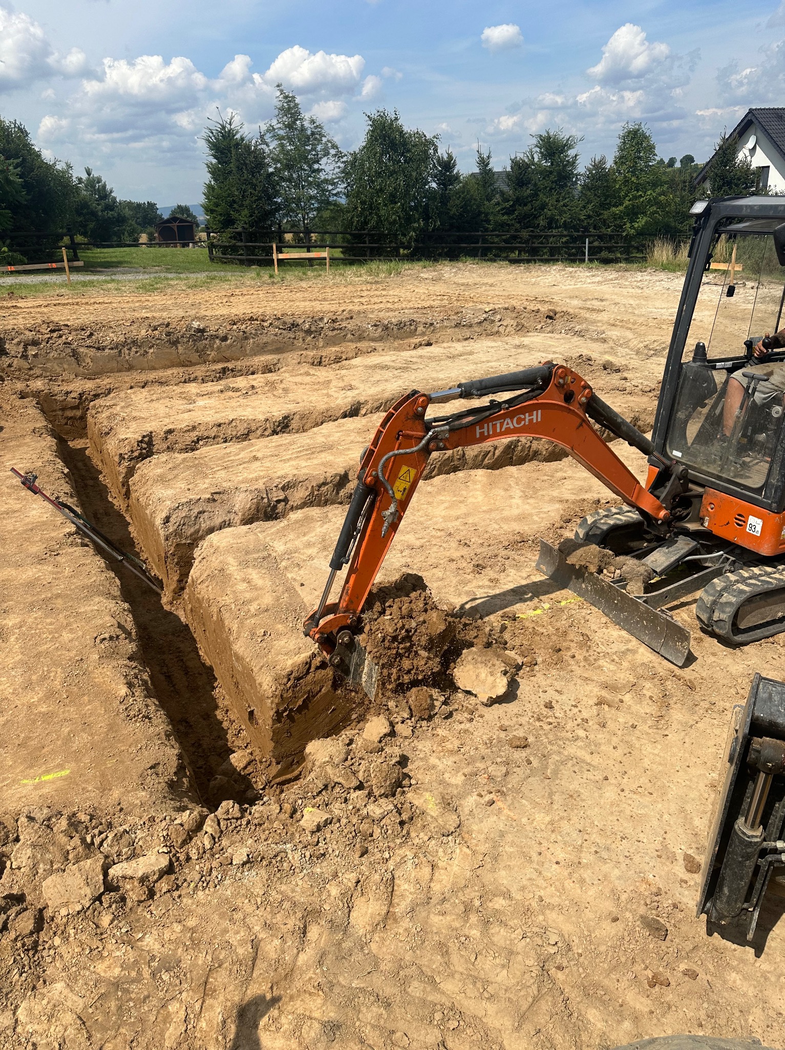 Pomarańczowa mini koparka Hitachi w trakcie wykopywania wąskiego rowu w ziemi, widoczne narzędzia pomiarowe w wykopie, przygotowanie pod fundament w słoneczny dzień.