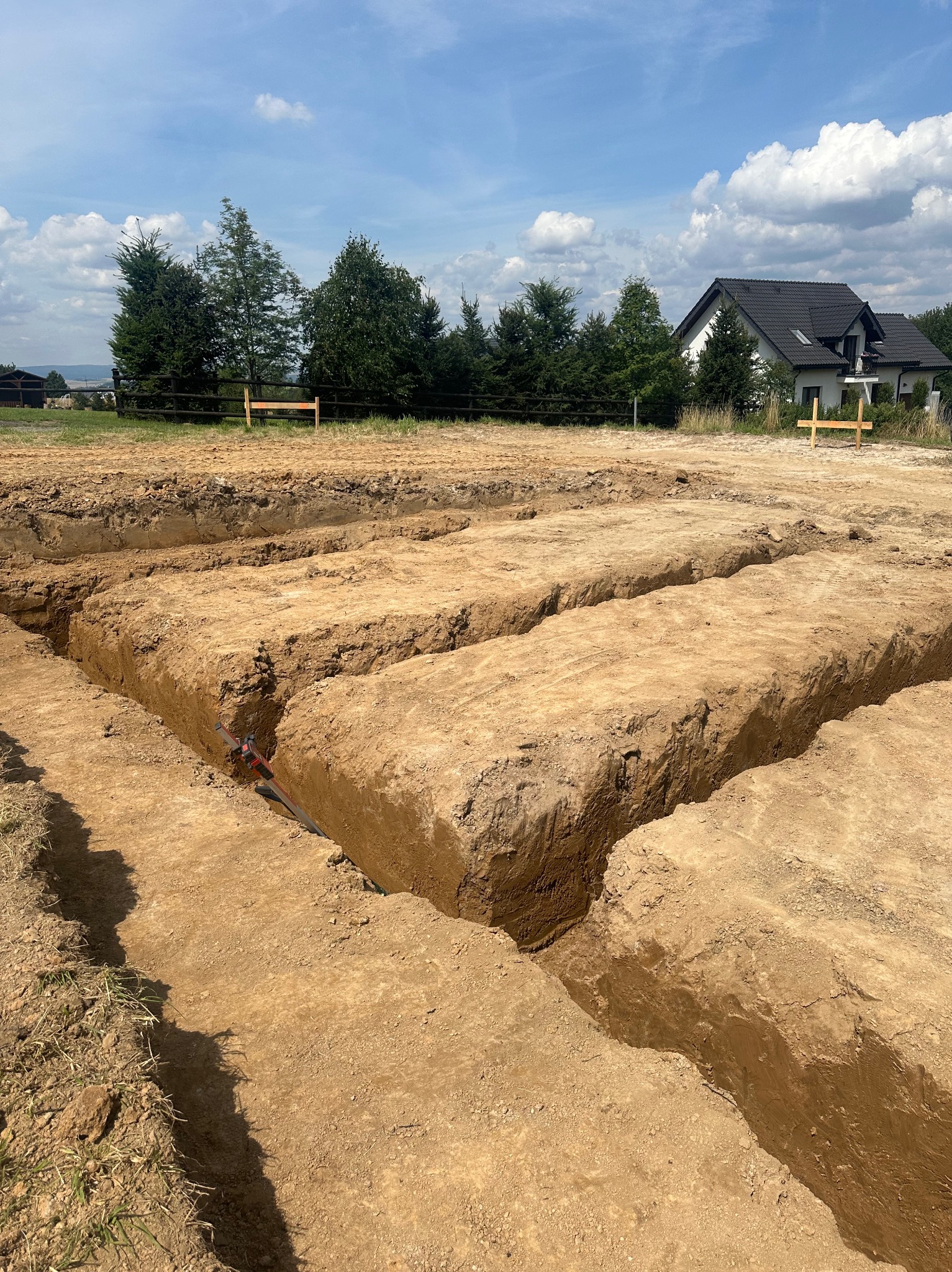 Wykopane rowy w ziemi pod fundamenty domu jednorodzinnego, widoczne instalacje, w tle dom i drzewa.