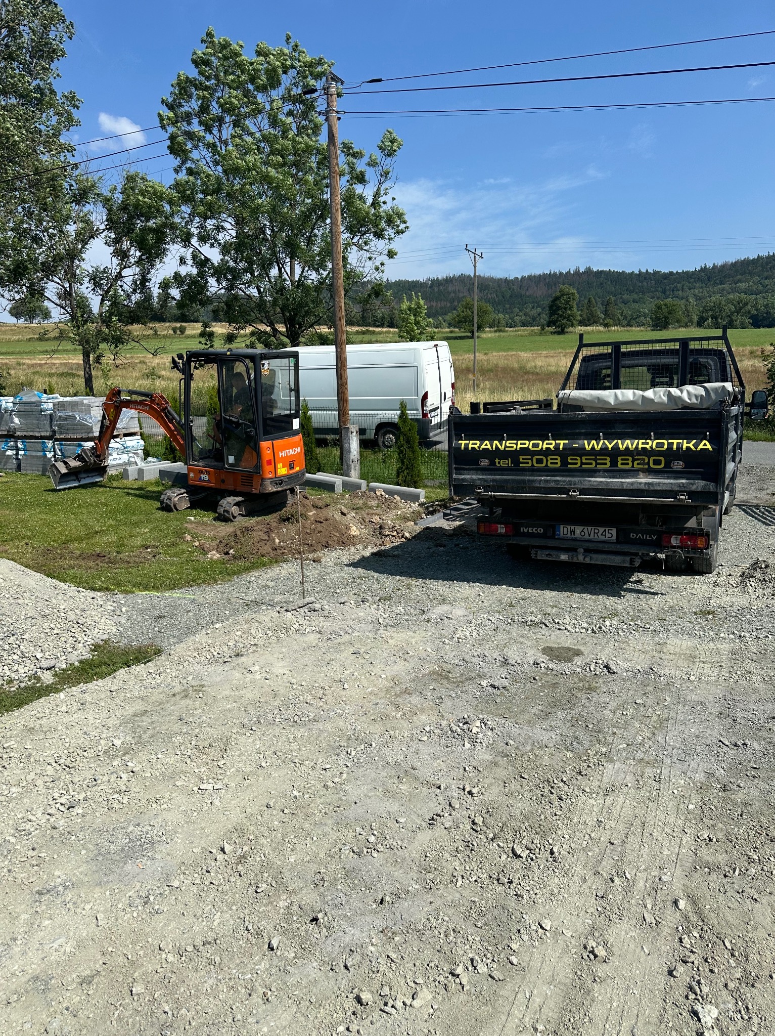 Pomarańczowa mini koparka Hitachi na trawniku obok palet z kostką brukową i częściowo ułożonych obrzeży, w tle biały bus i czarna wywrotka z numerem telefonu na skrzyni ładunkowej.