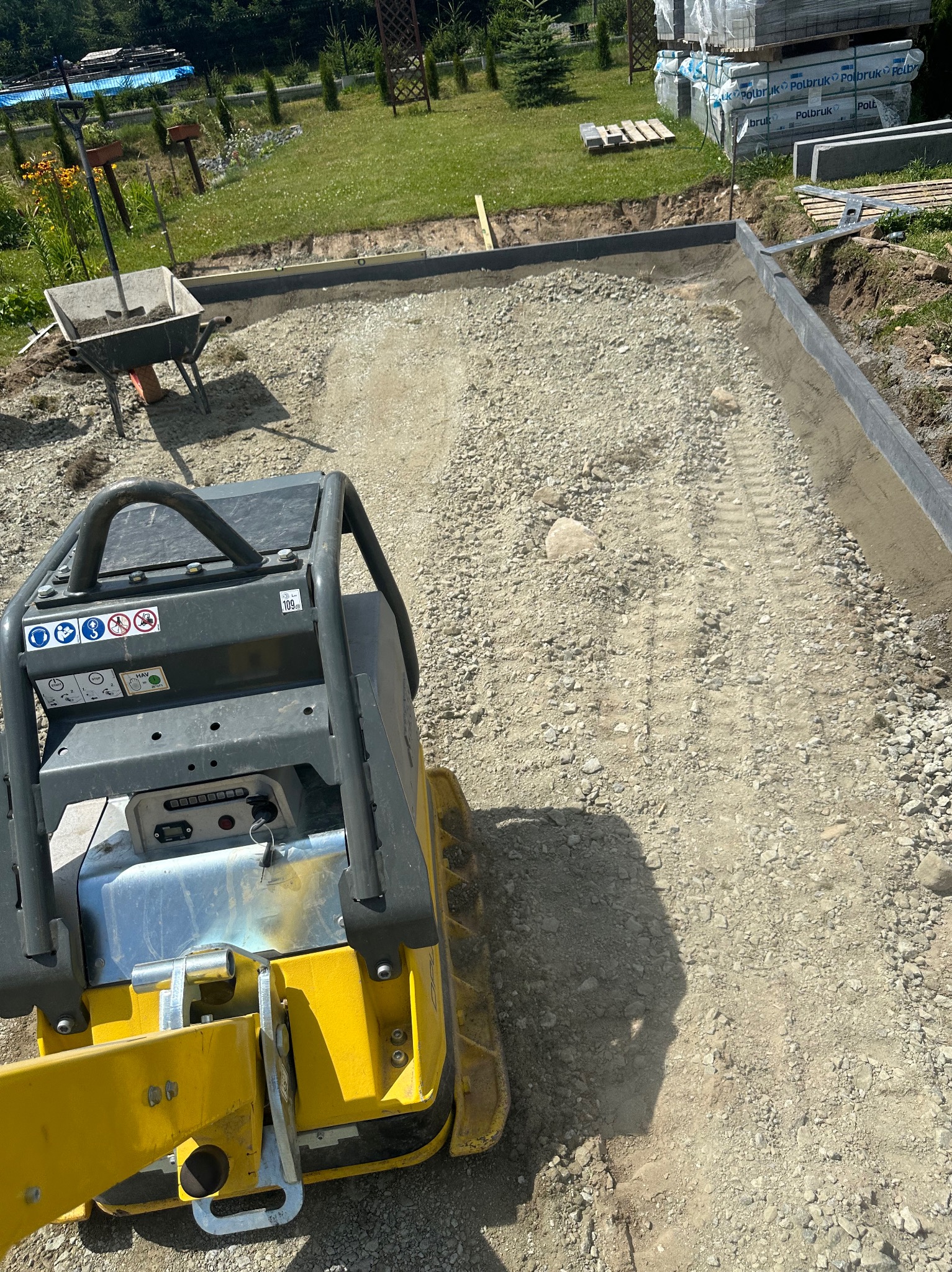 Żółta zagęszczarka wibrująca na podsypce pod przyszłą nawierzchnię, obrzeża ułożone, w tle palety kostki brukowej.