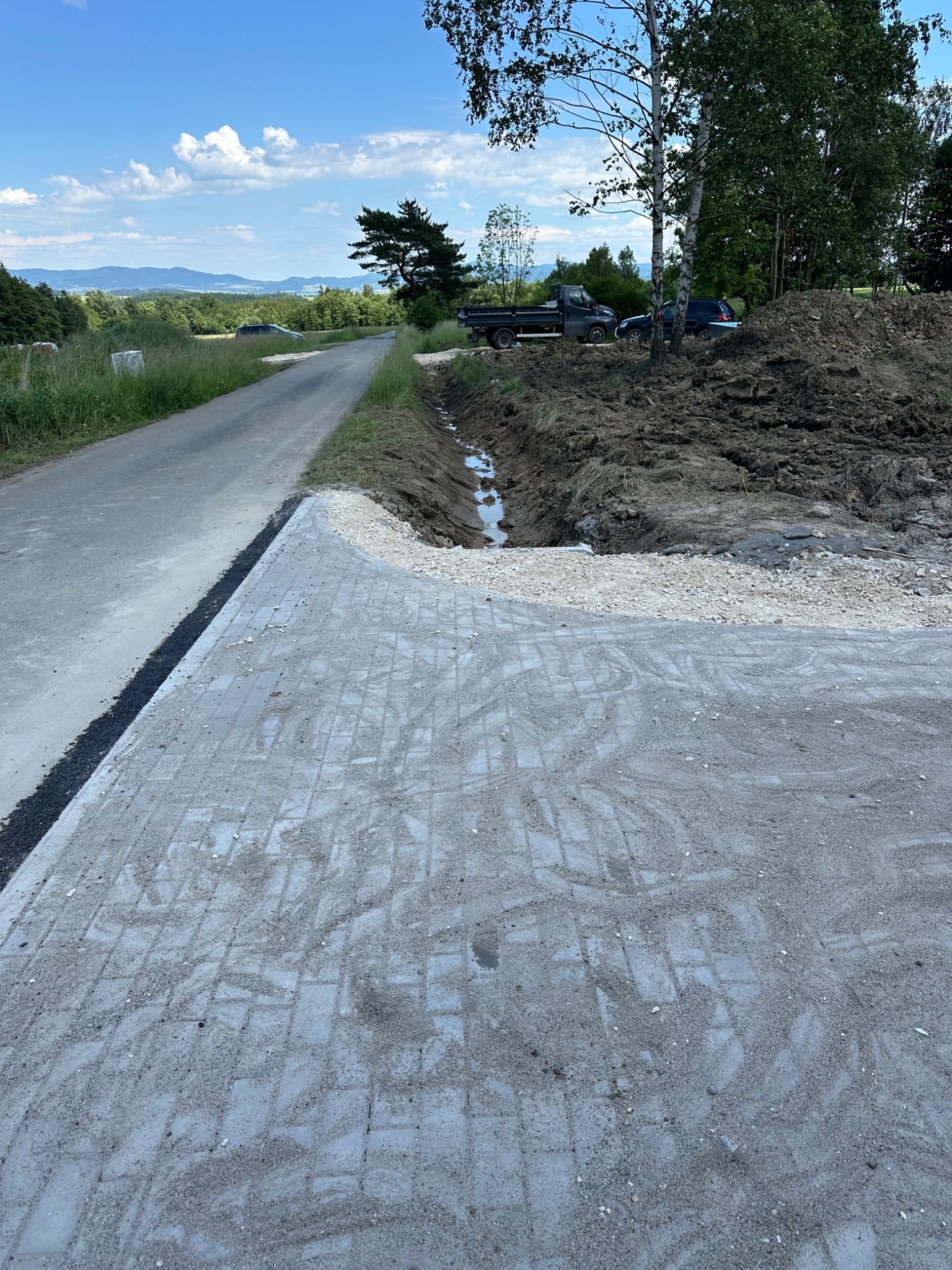 Nowa nawierzchnia z kostki brukowej sąsiadująca z asfaltową drogą, w tle wykop z wodą i sterta ziemi oraz ciężarówka i samochód osobowy