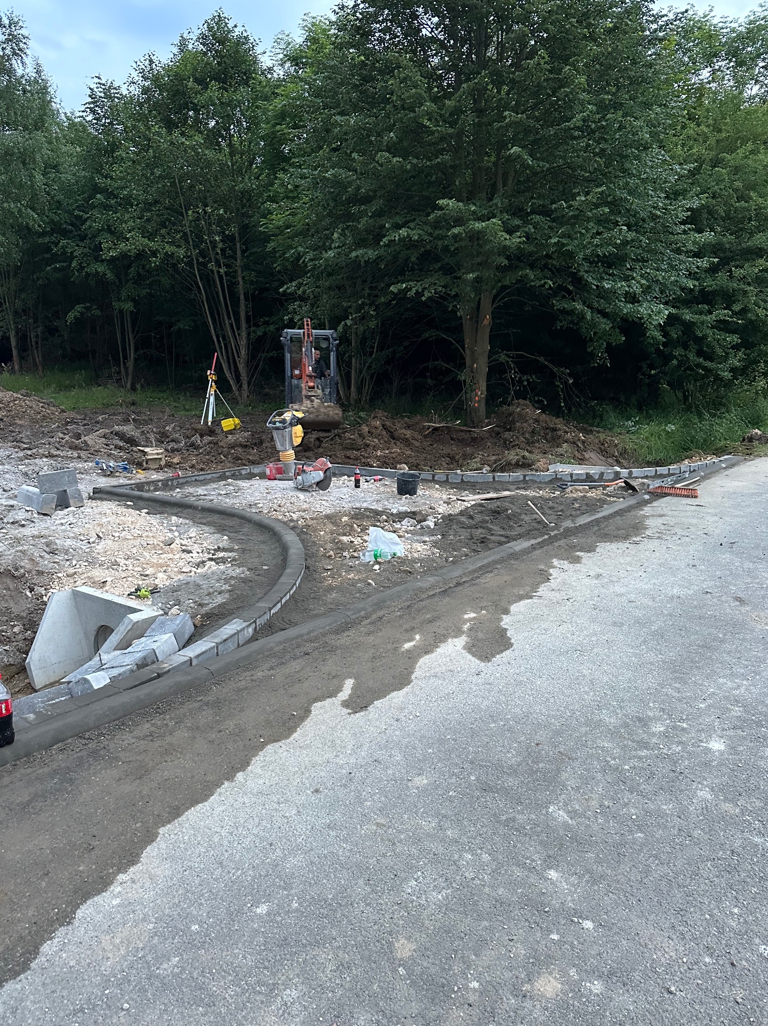 Budowa drogi z obrzeżami betonowymi, widoczny minikoparka, niwelator i zagęszczarka, w tle zalesiony teren.