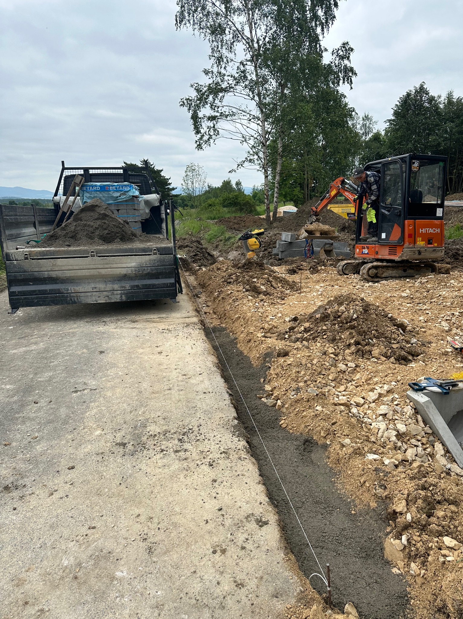 Przygotowanie terenu pod budowę; widoczny wykop z wylanym betonem, mini koparka Hitachi, ubijarka, paleta z kostką brukową na ciężarówce, tło z drzewami.