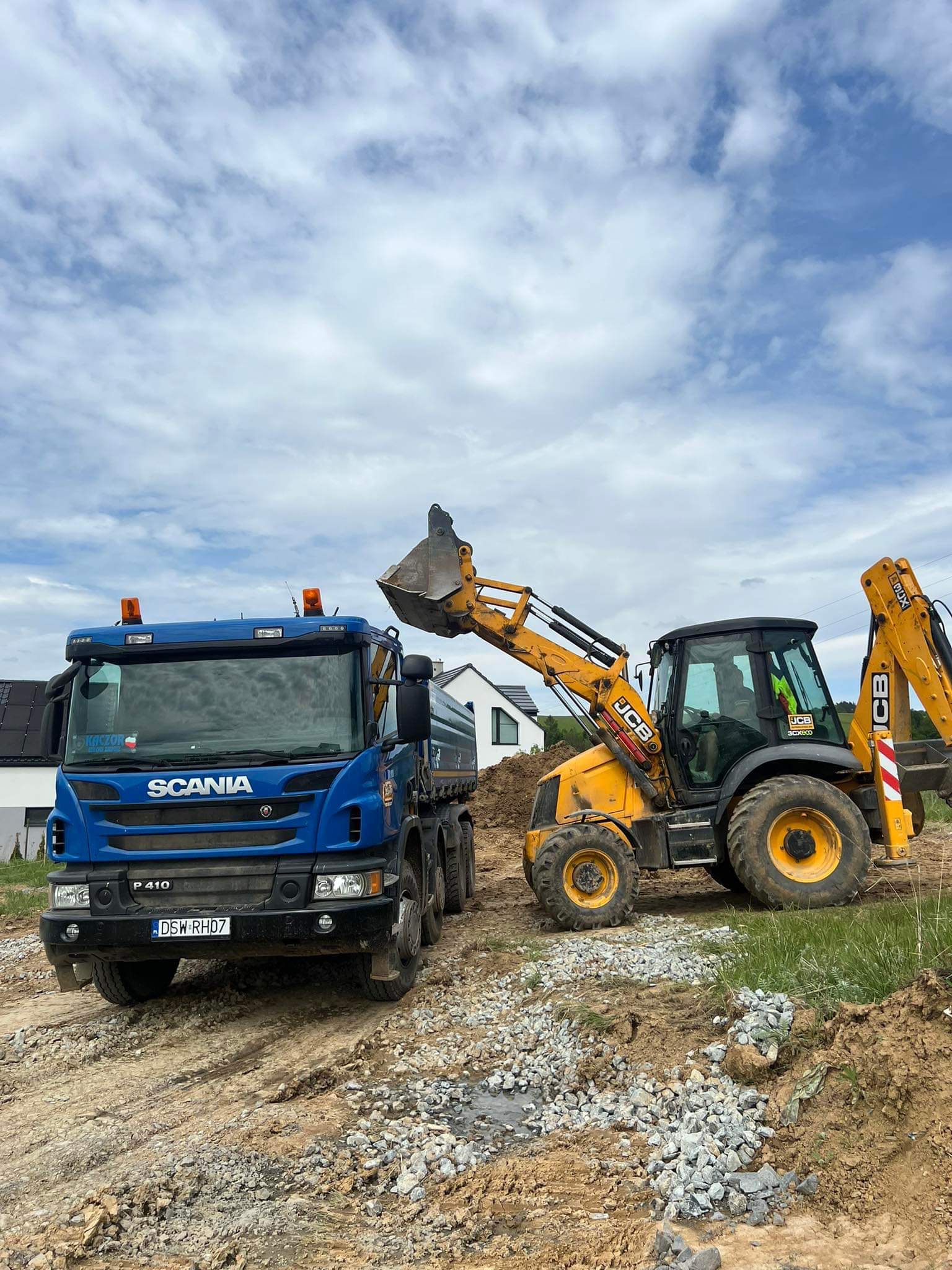Niebieska ciężarówka Scania i żółta koparko-ładowarka JCB na placu budowy z gruzem, w tle dom jednorodzinny i błękitne niebo z chmurami.