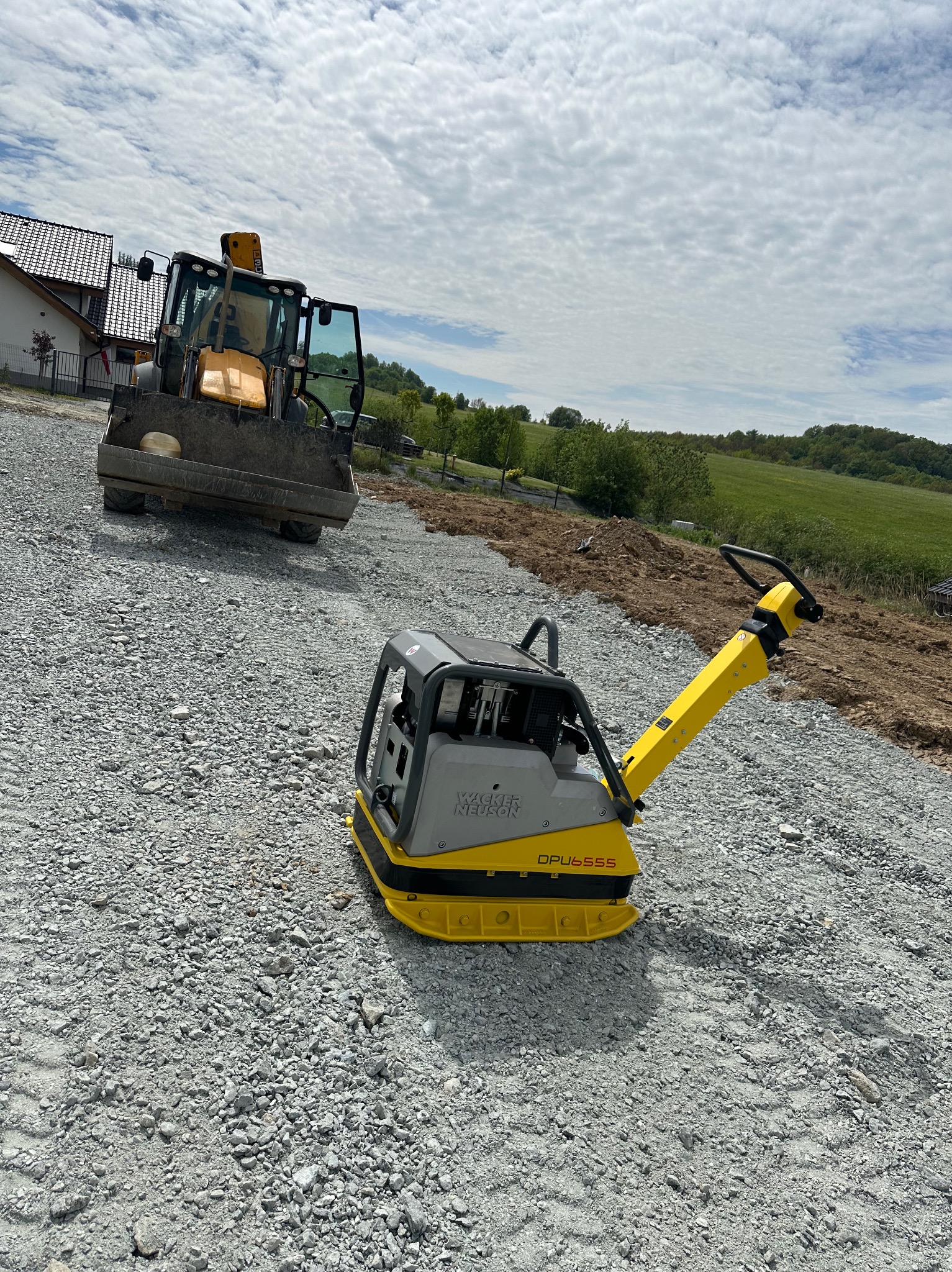 Żółto-szara zagęszczarka Wacker Neuson DPU6555 na podsypce z tłucznia, w tle koparko-ładowarka JCB i teren przygotowany pod budowę.