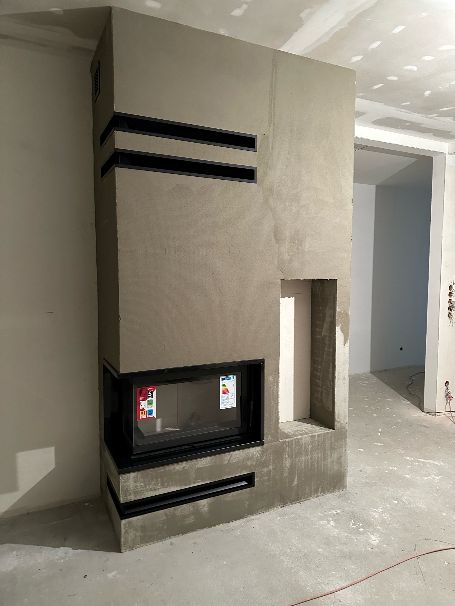 Surowy, szary obudowany wkład kominkowy z czarnymi, poziomymi szczelinami wentylacyjnymi w trakcie prac wykończeniowych, widoczne niedoskonałości tynku i elementy instalacji elektrycznej w tle.