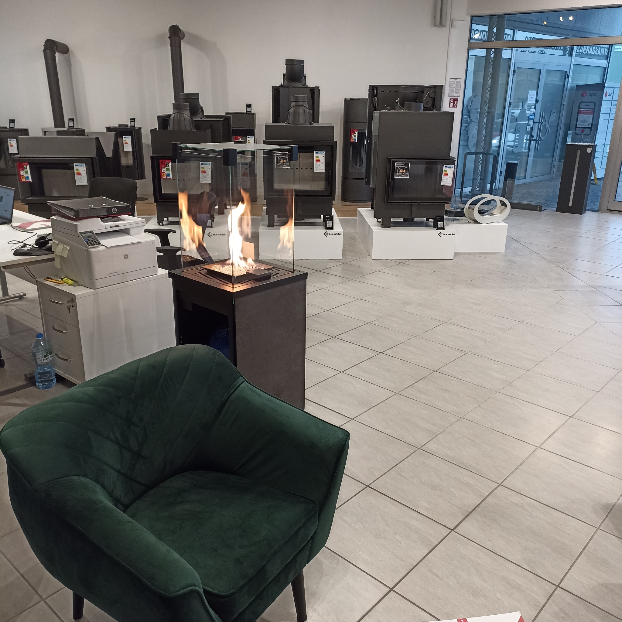 Ekspozycja kominków marki Kratki.pl w salonie: biokominek z płomieniami za szybą, obok szereg pieców kominkowych na ekspozycyjnych podestach, w tle widoczne wejście do salonu.