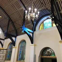 Wnętrze kościoła w trakcie odnawiania, widoczne rusztowania, odnowione witraże w łukowych oknach, pomalowane na biało ściany z elementami dekoracyjnymi w kolorze złotym oraz drewniany sufit...