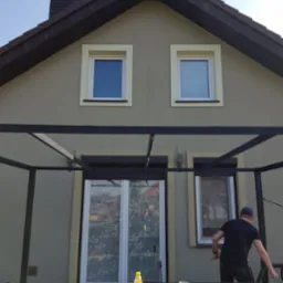 Malowanie elewacji domu jednorodzinnego w trakcie realizacji, widoczny malarz z pędzlem, wiadro z farbą, st&oacute;ł z narzędziami i akcesoriami malarskimi oraz fragment budynku z oknami i pergolą.