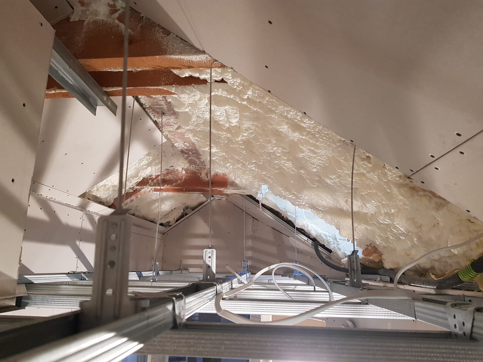 Poddasze w trakcie ocieplania pianą PUR, widoczne metalowe profile do montażu płyt kartonowo-gipsowych, przewody elektryczne i fragment drewnianej konstrukcji dachu.