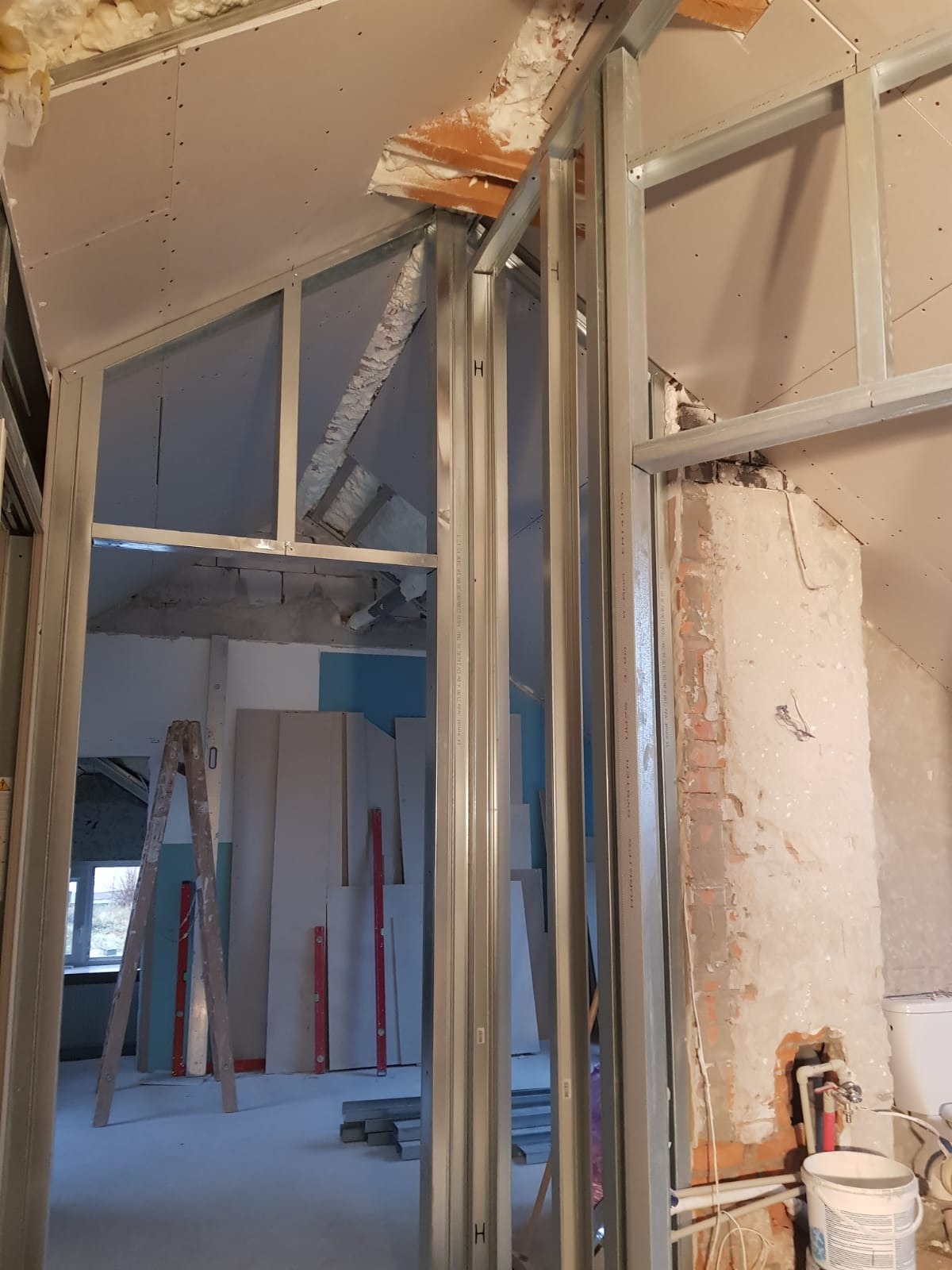 Montaż metalowej konstrukcji ścian działowych na poddaszu, widoczne płyty karton-gips, drabina oparta o ścianę, instalacja hydrauliczna w tle, fragment odsłoniętej cegły.