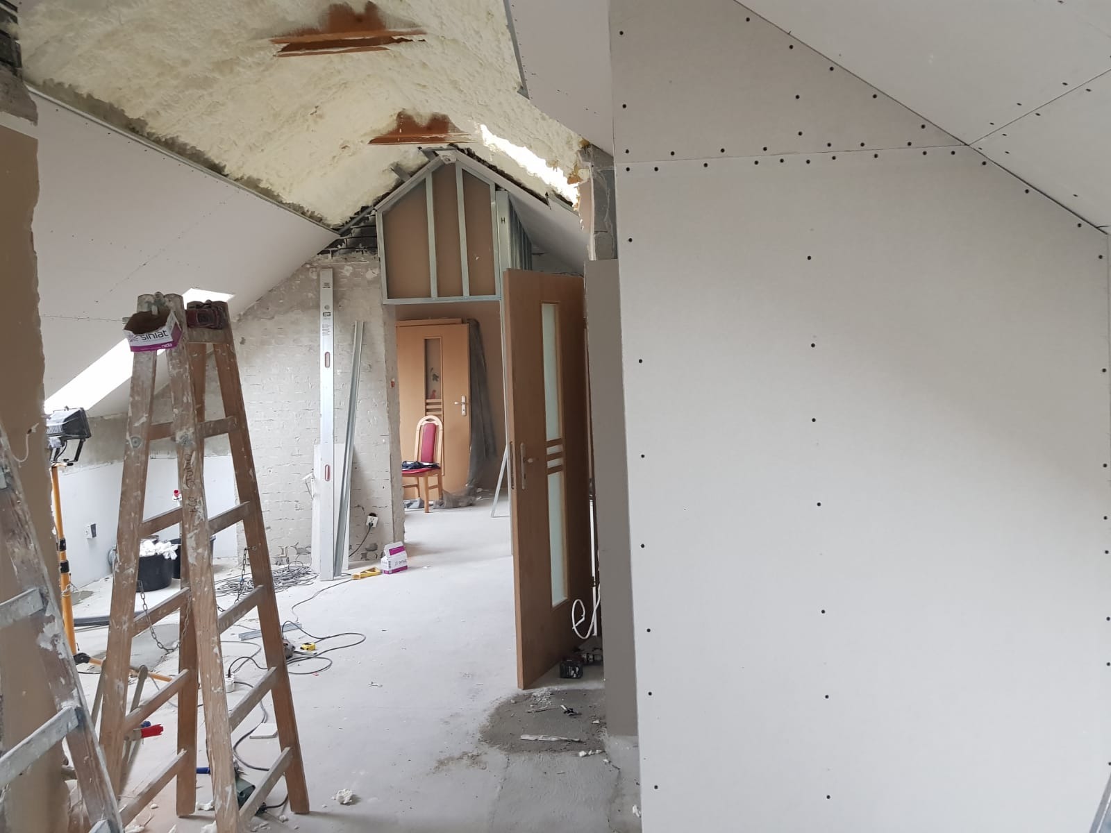 Poddasze w trakcie adaptacji: widoczne płyty kartonowo-gipsowe na skosach dachu, izolacja pianką montażową, metalowe profile konstrukcyjne i drewniana drabina. W tle otwarte drzwi do kolejnego...