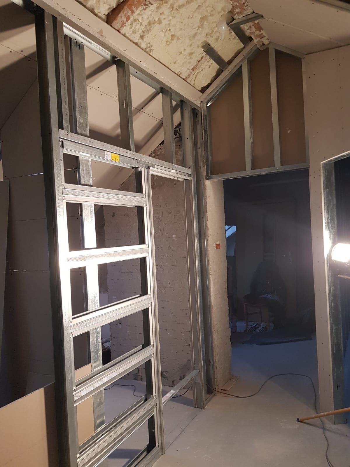 Montaż metalowej konstrukcji szkieletowej na poddaszu, widoczne profile ścienne i sufitowe, częściowo obłożone płytami gipsowo-kartonowymi, surowa cegła w tle.