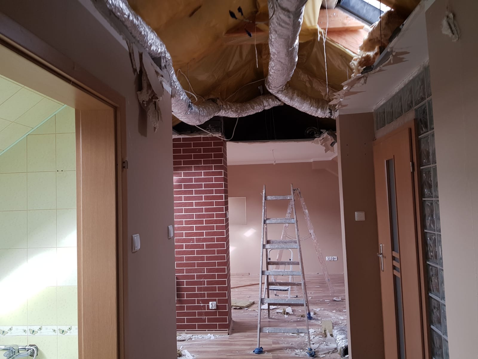 Wnętrze w trakcie demontażu sufitu podwieszanego, widoczne fragmenty instalacji wentylacyjnej, drabina i pozostałości po pracach na podłodze, z widokiem na pomieszczenie z ceglaną ścianą.