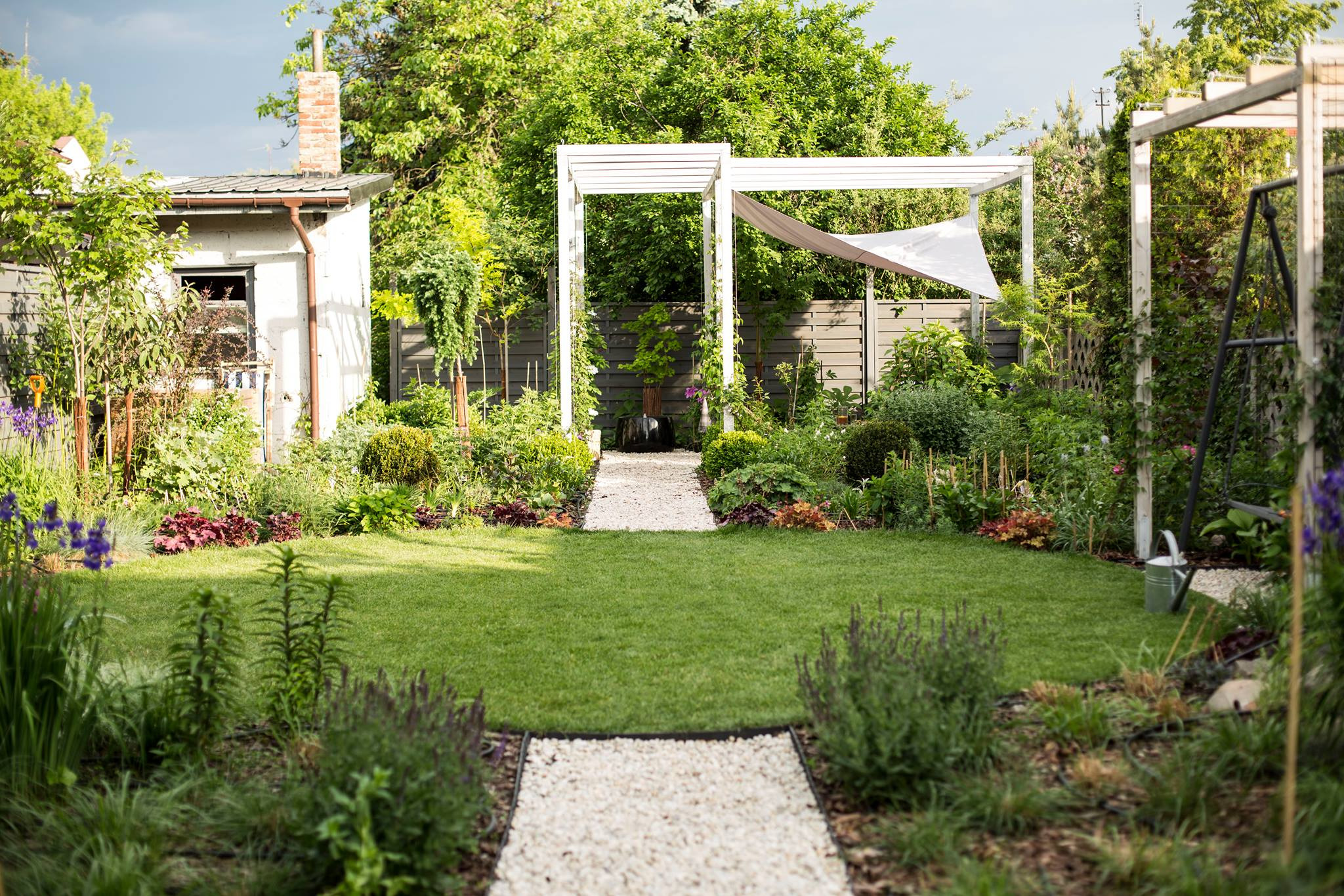 Utrzymany ogród z równo przystrzyżonym trawnikiem, ścieżką z jasnego żwiru prowadzącą do białej pergoli z zasłoną przeciwsłoneczną, na tyłach drewniany płot i bujna roślinność, w tle mały budynek...
