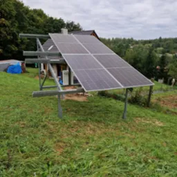 Panele słoneczne zamontowane na metalowej konstrukcji na trawiastym zboczu, w tle dom z brązowym dachem i drzewa pod pochmurnym niebem.