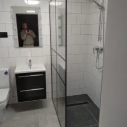 Nowoczesna łazienka z kabiną prysznicową typu walk-in z czarnymi profilami, umywalką z szafką w kolorze czarnym, toaletą i lustrem, w którym odbija się osoba robiąca zdjęcie.