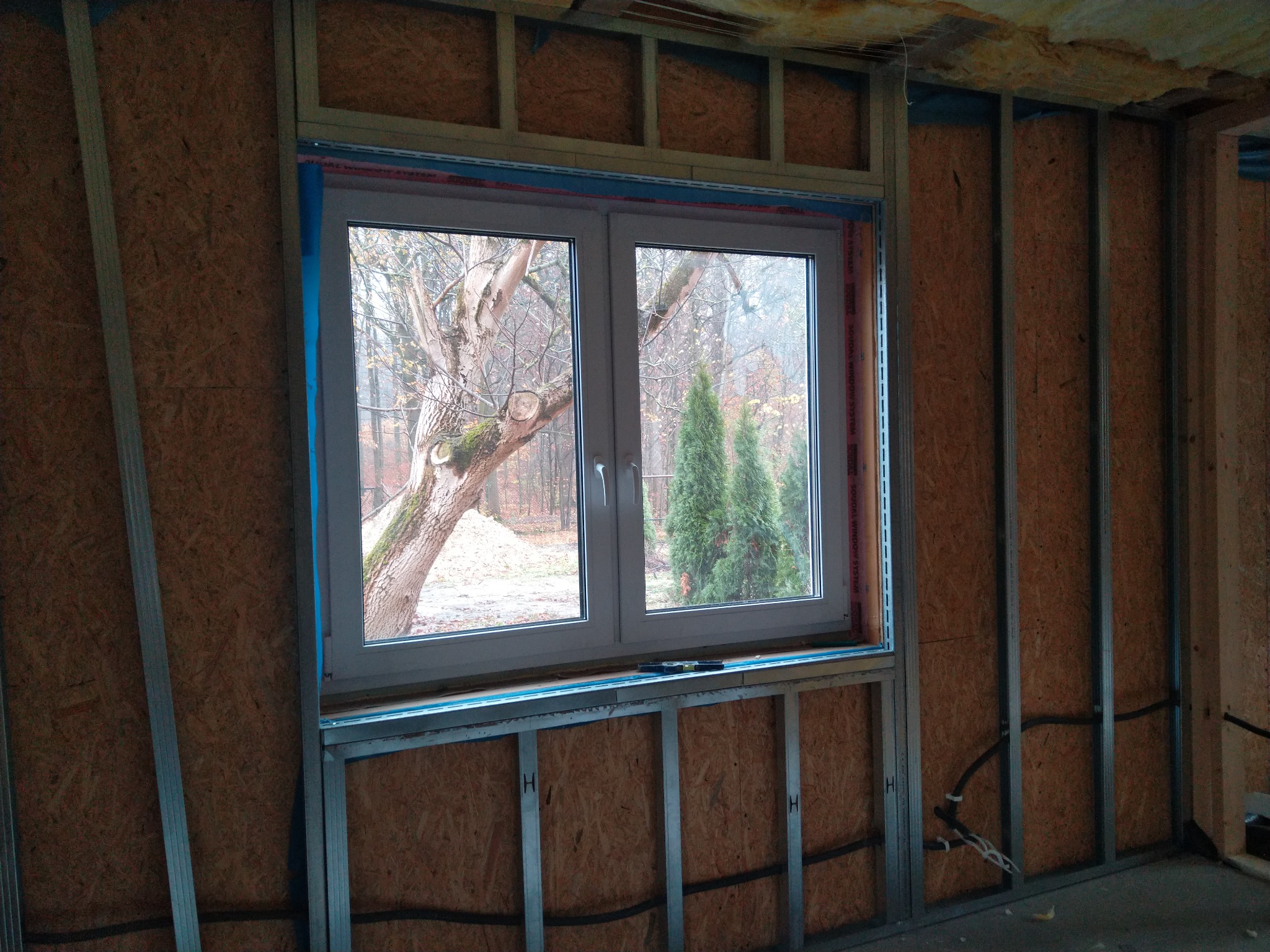 Montaż okna PCV w konstrukcji szkieletowej z płyt OSB, widoczne profile metalowe i instalacja elektryczna w ścianie.