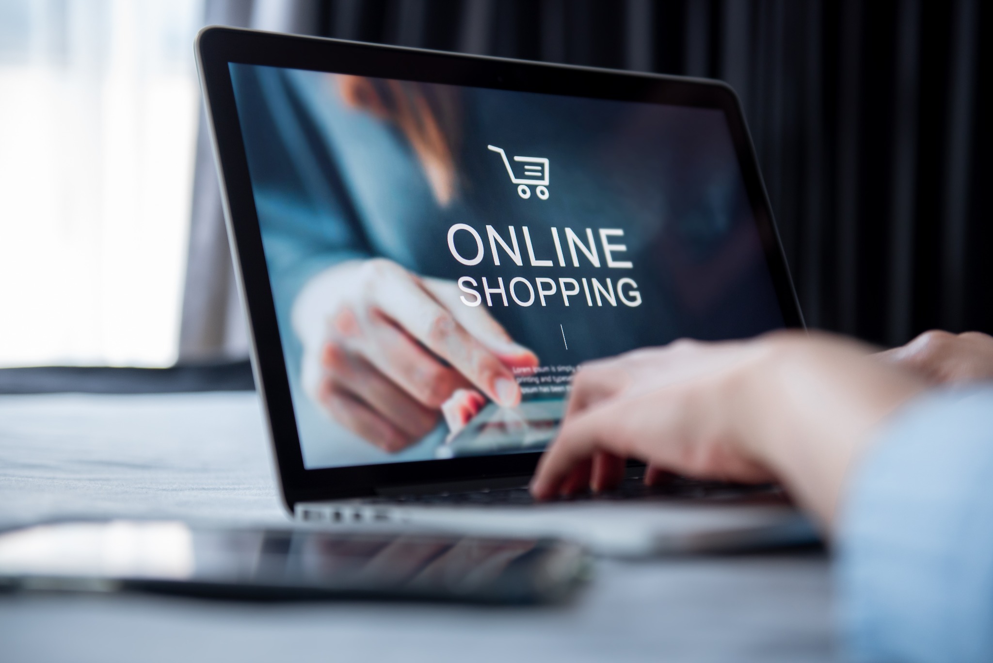 Laptop z ekranem wyświetlającym napis ONLINE SHOPPING i ikonę wózka sklepowego, dłonie użytkownika w tle, telefon na pierwszym planie.