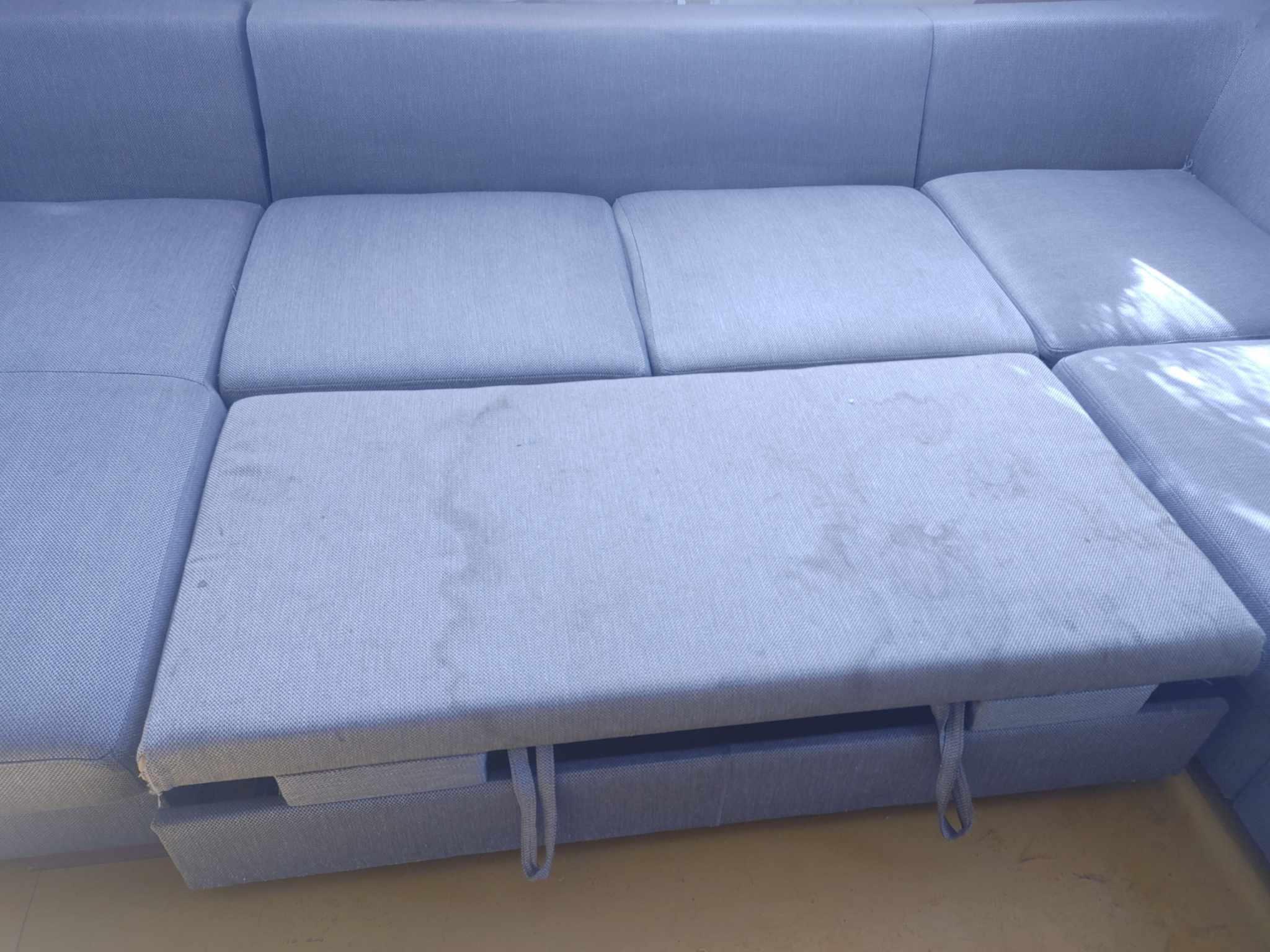 Szara, modułowa kanapa z widocznymi plamami na jednym z siedzisk, które jest lekko wysunięte, ukazując konstrukcję pod spodem.