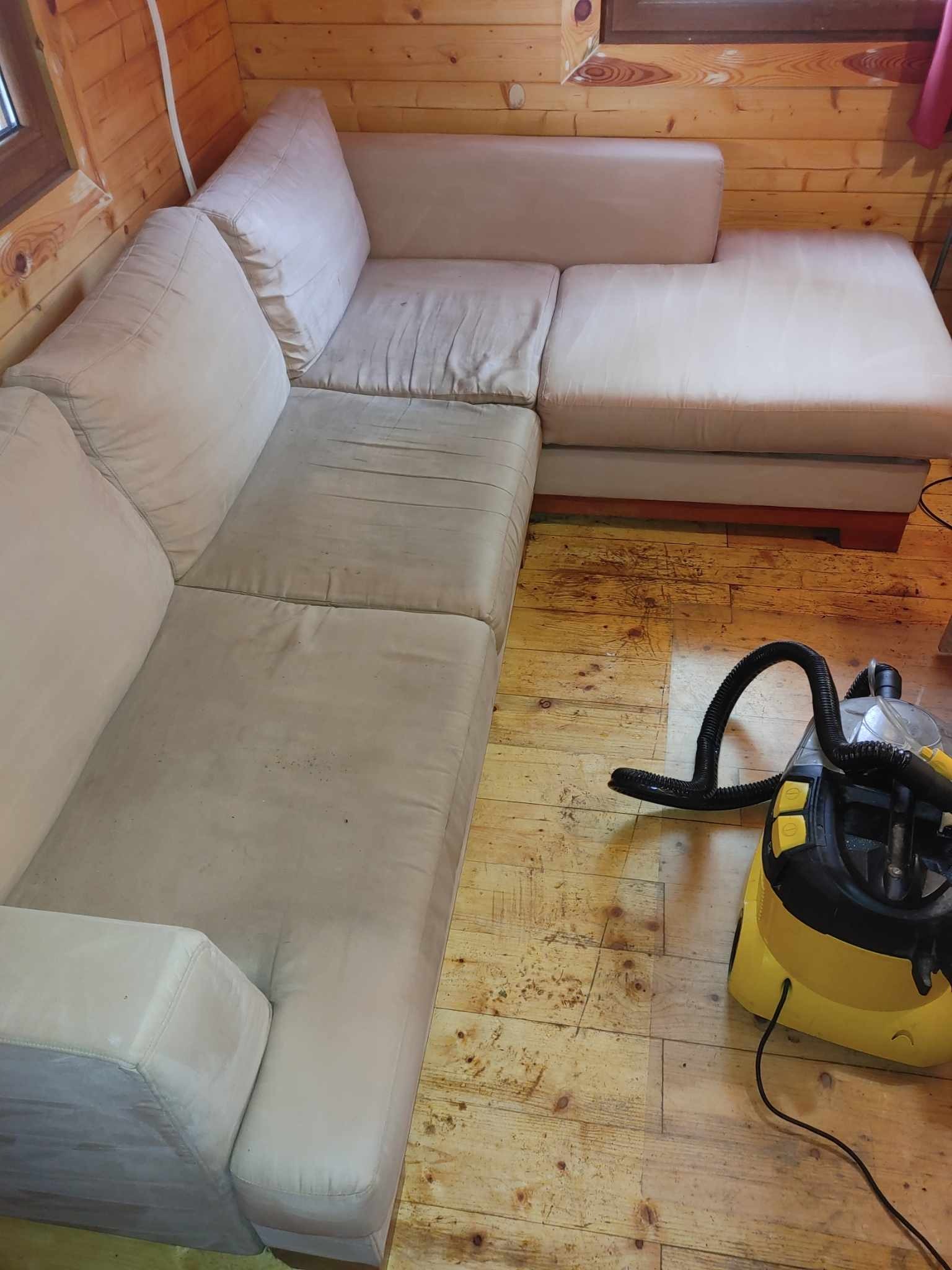 Narożnik tapicerowany z widocznymi plamami poddawany czyszczeniu przy użyciu żółtego odkurzacza piorącego na drewnianej podłodze w domku z bali.