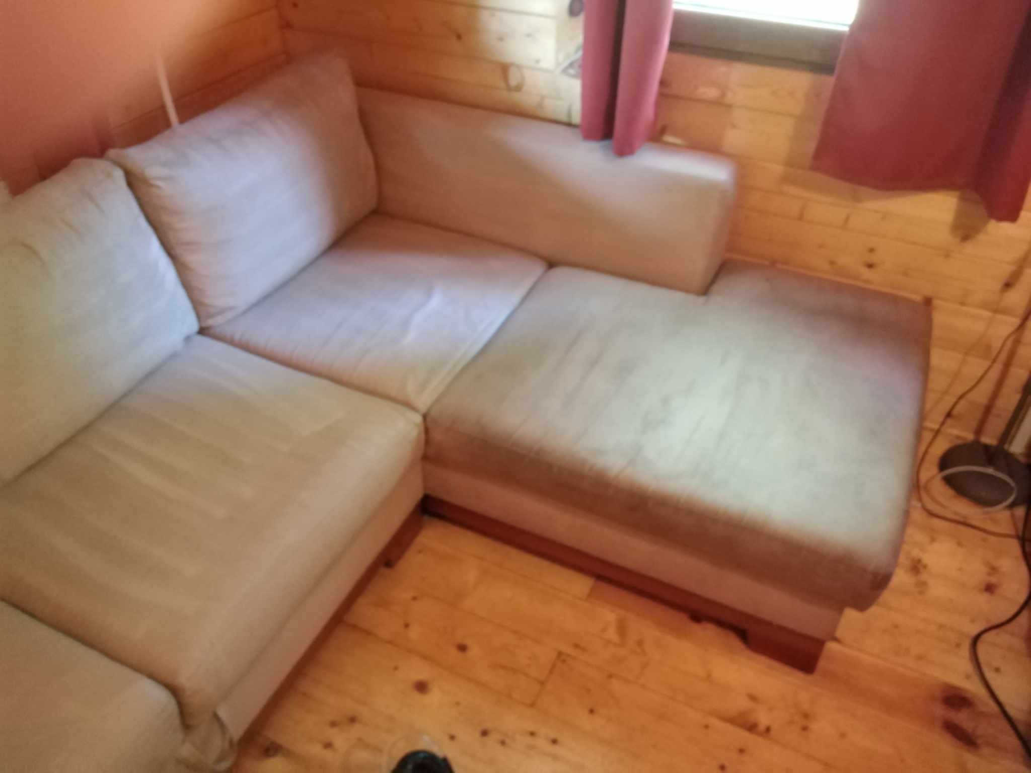 Jasna, narożna kanapa z wyraźnymi śladami użytkowania na siedziskach, sfotografowana w drewnianym wnętrzu z widocznymi czerwonymi zasłonami przy oknie.