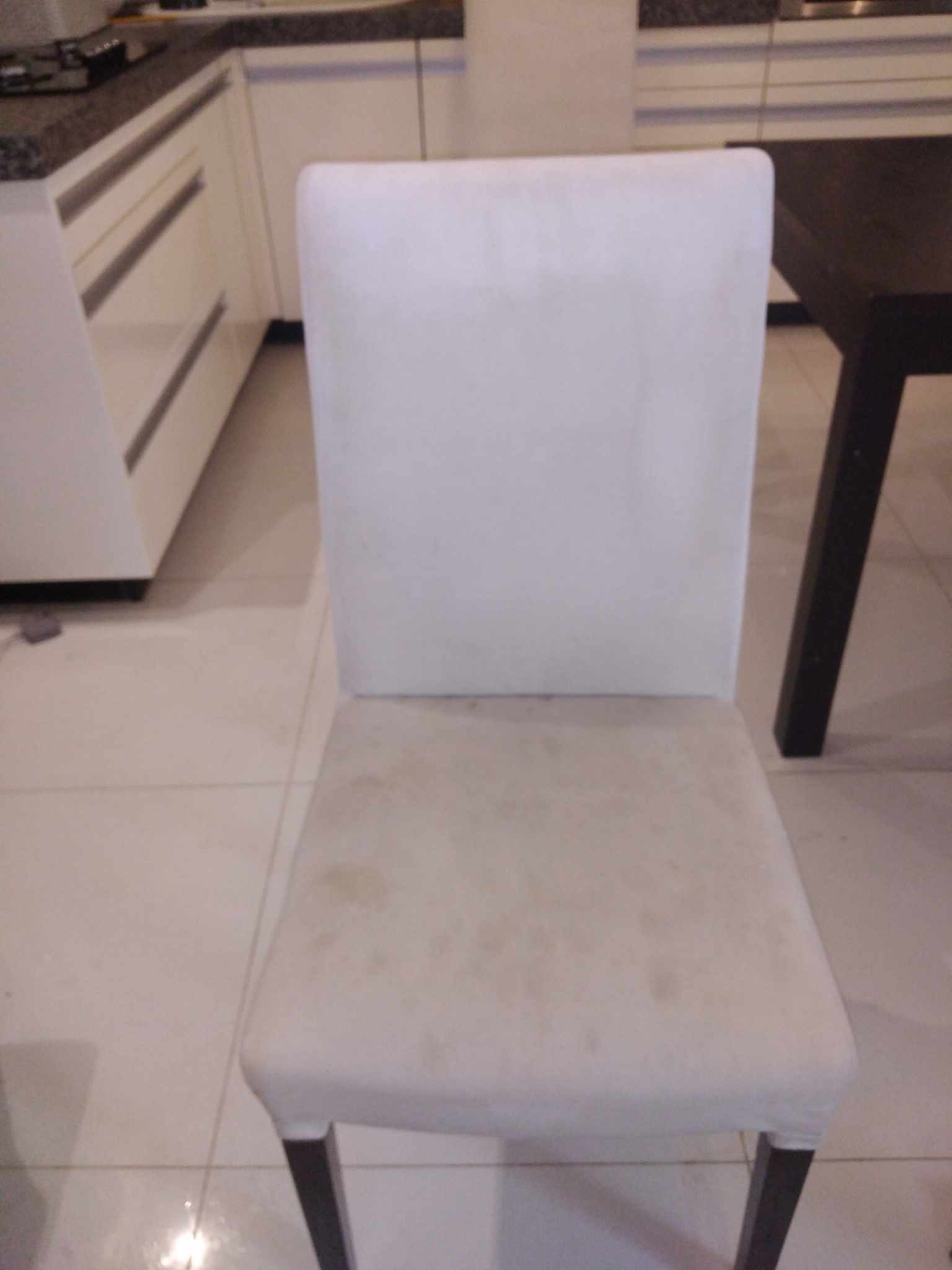 Białe krzesło tapicerowane z widocznymi plamami na siedzisku, stojące na kafelkach w kuchni. W tle widoczny fragment blatu i białych szafek kuchennych.