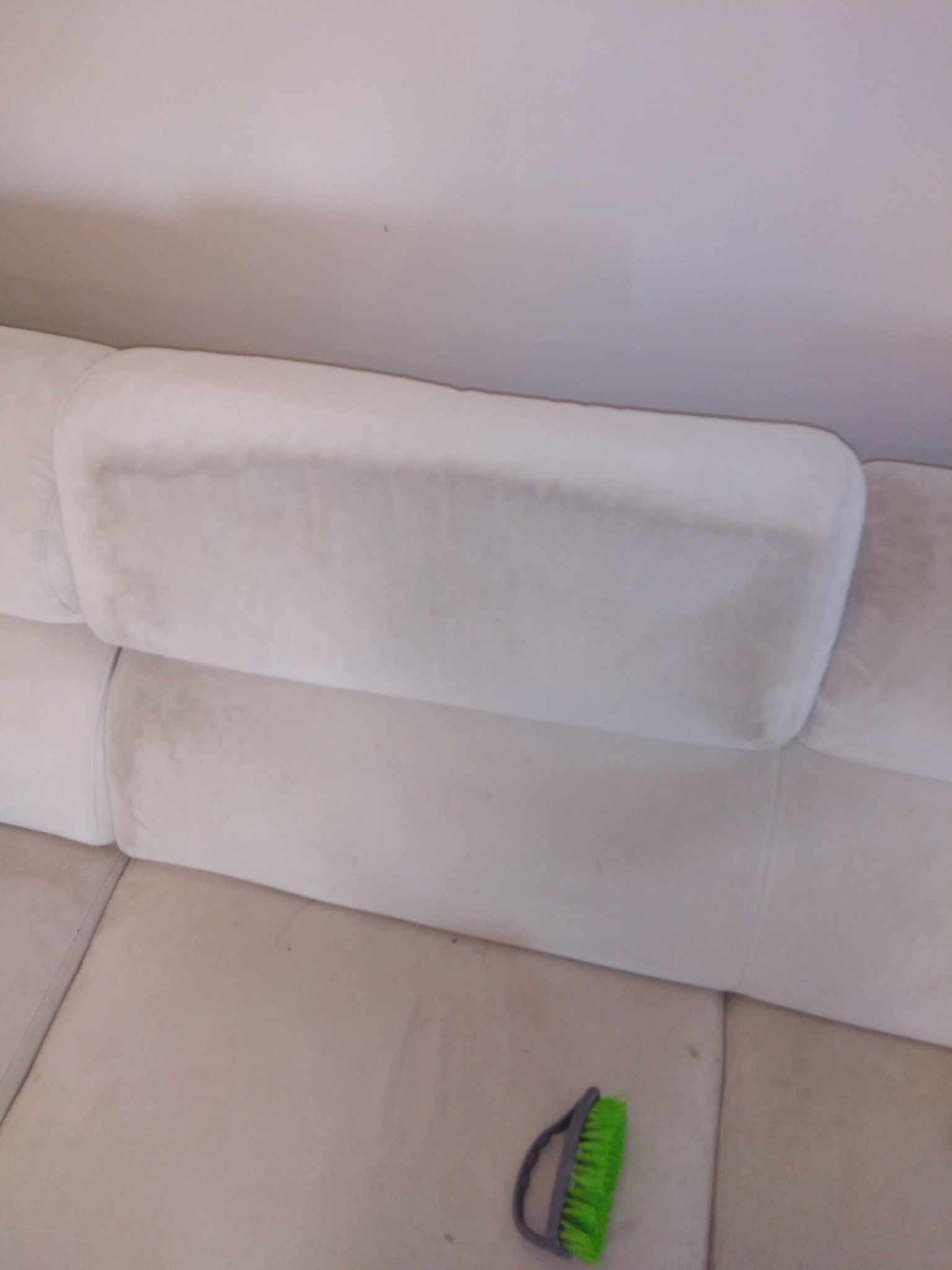 Fragment sofy w jasnym odcieniu z widocznymi plamami i szczotką do czyszczenia na dole kadru.