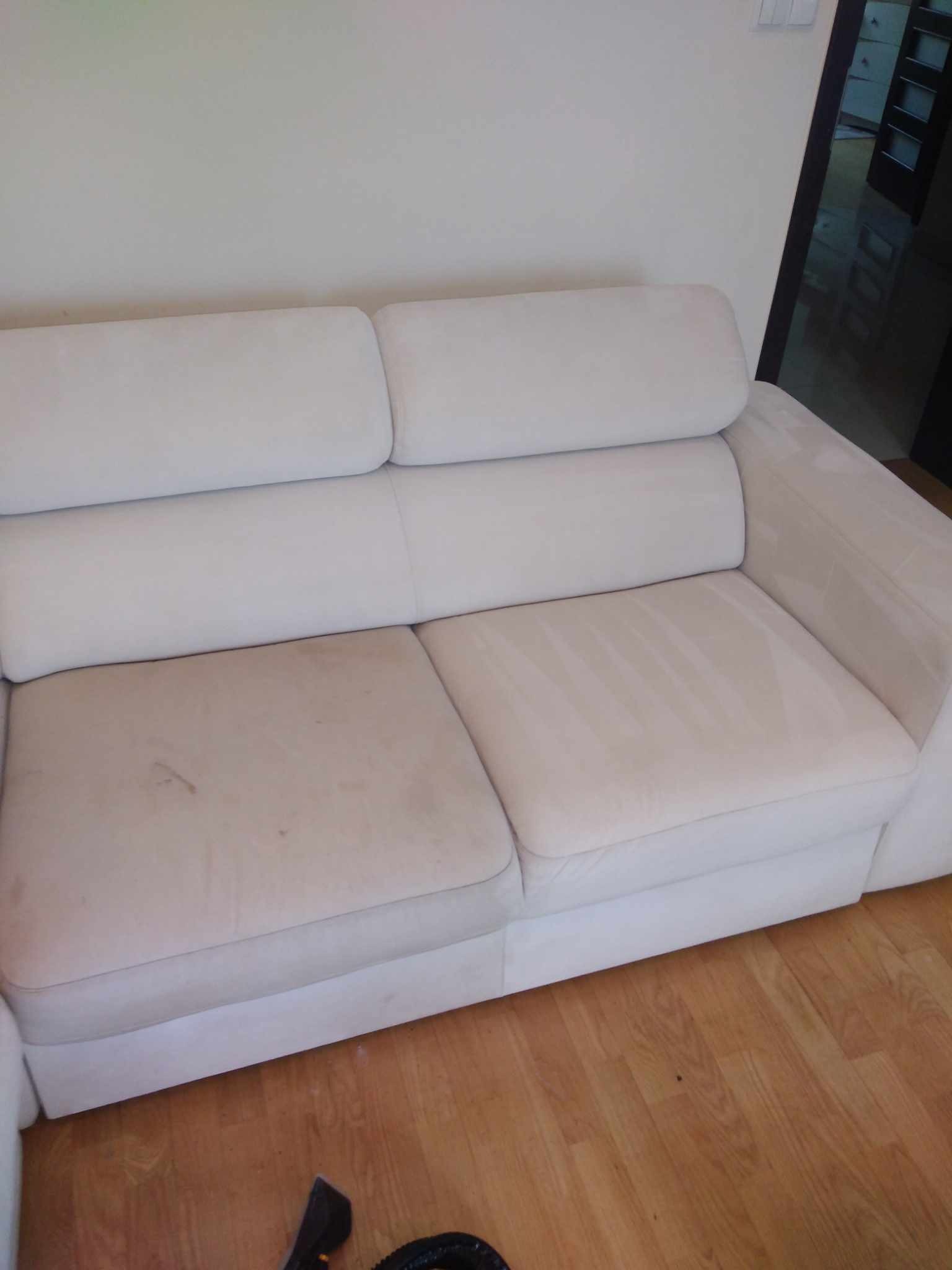 Dwusiedzeniowa, jasna kanapa z widocznymi plamami na jednym siedzisku, częściowo wyczyszczona z widocznymi śladami po czyszczeniu, fragment podłogi z widocznym urządzeniem do czyszczenia.