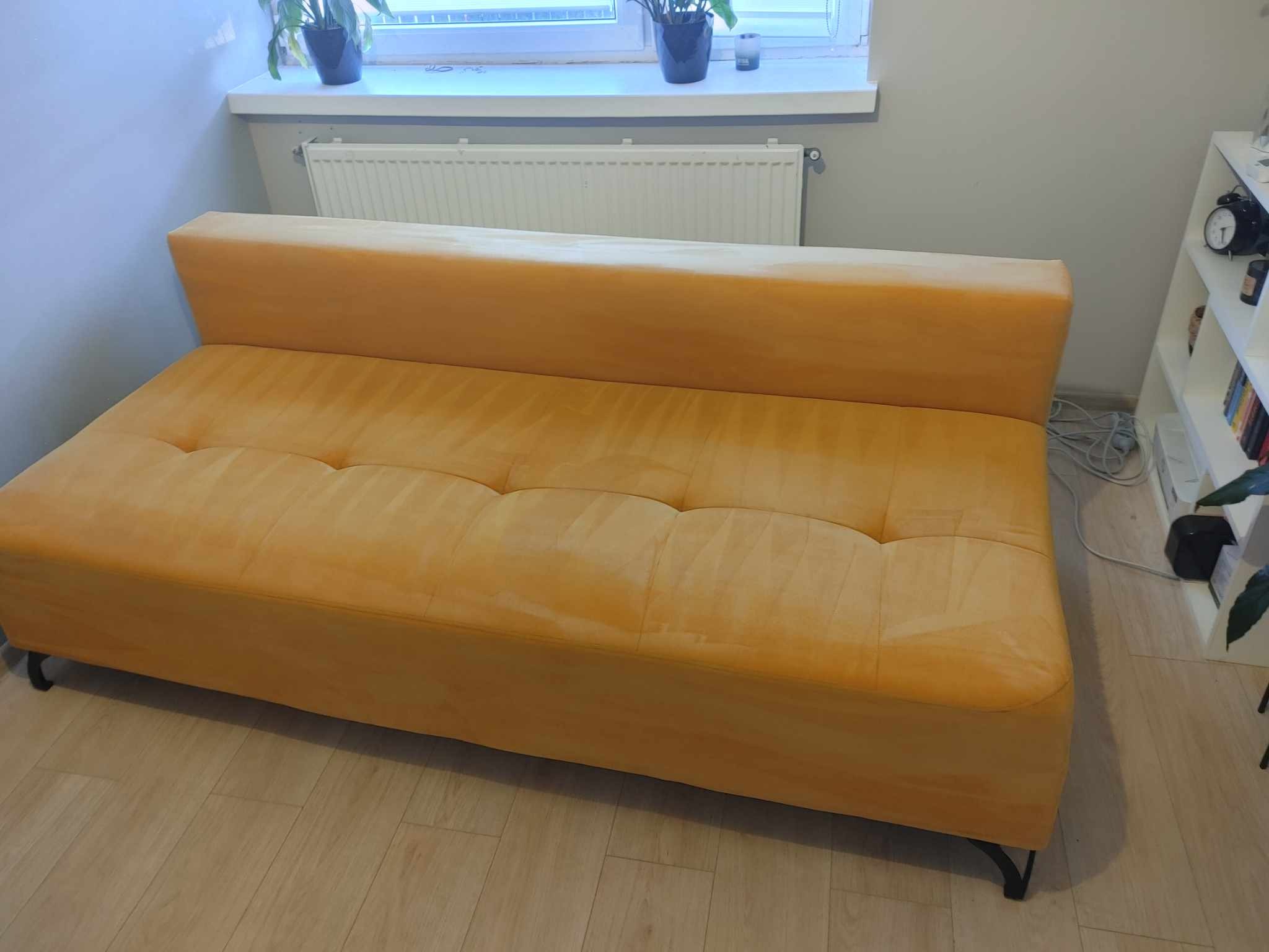 Żółta, welurowa kanapa z niskim oparciem i pikowanymi poduchami na drewnianej podłodze, widok z boku, w tle biały grzejnik i półka z dekoracjami.