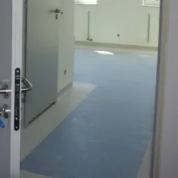 Widok zza uchylonych drzwi na jasne pomieszczenie z dwukolorową podłogą z tworzywa sztucznego: błękitna powierzchnia przechodzi w szarą w pobliżu ściany. Na ścianach widoczne białe listwy...