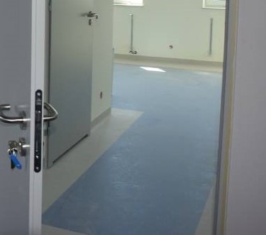 Widok zza uchylonych drzwi na jasne pomieszczenie z dwukolorową podłogą z tworzywa sztucznego: błękitna powierzchnia przechodzi w szarą w pobliżu ściany. Na ścianach widoczne białe listwy...