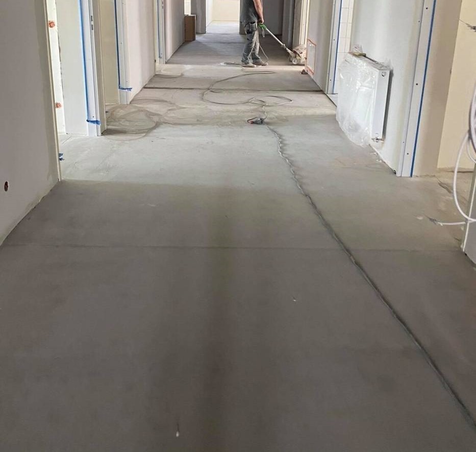 Wyrównywanie podłoża w długim korytarzu przed położeniem nowej podłogi, widoczny pracownik z szlifierką do betonu, zabezpieczone drzwi taśmą malarską oraz zakryty grzejnik.