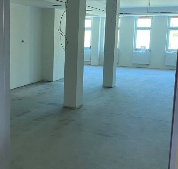 Surowy, betonowy podkład podłogowy w przestronnym, jasnym pomieszczeniu biurowym z białymi kolumnami i grzejnikami pod oknami, przygotowany do dalszych prac wykończeniowych.