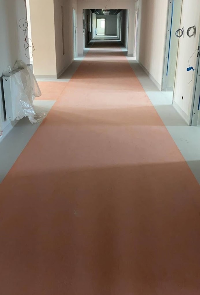Długi korytarz w budynku w trakcie remontu, z nową, jednolitą, pomarańczową powierzchnią podłogową, prawdopodobnie z wykładziny, z widocznymi instalacjami i grzejnikiem zabezpieczonym folią.