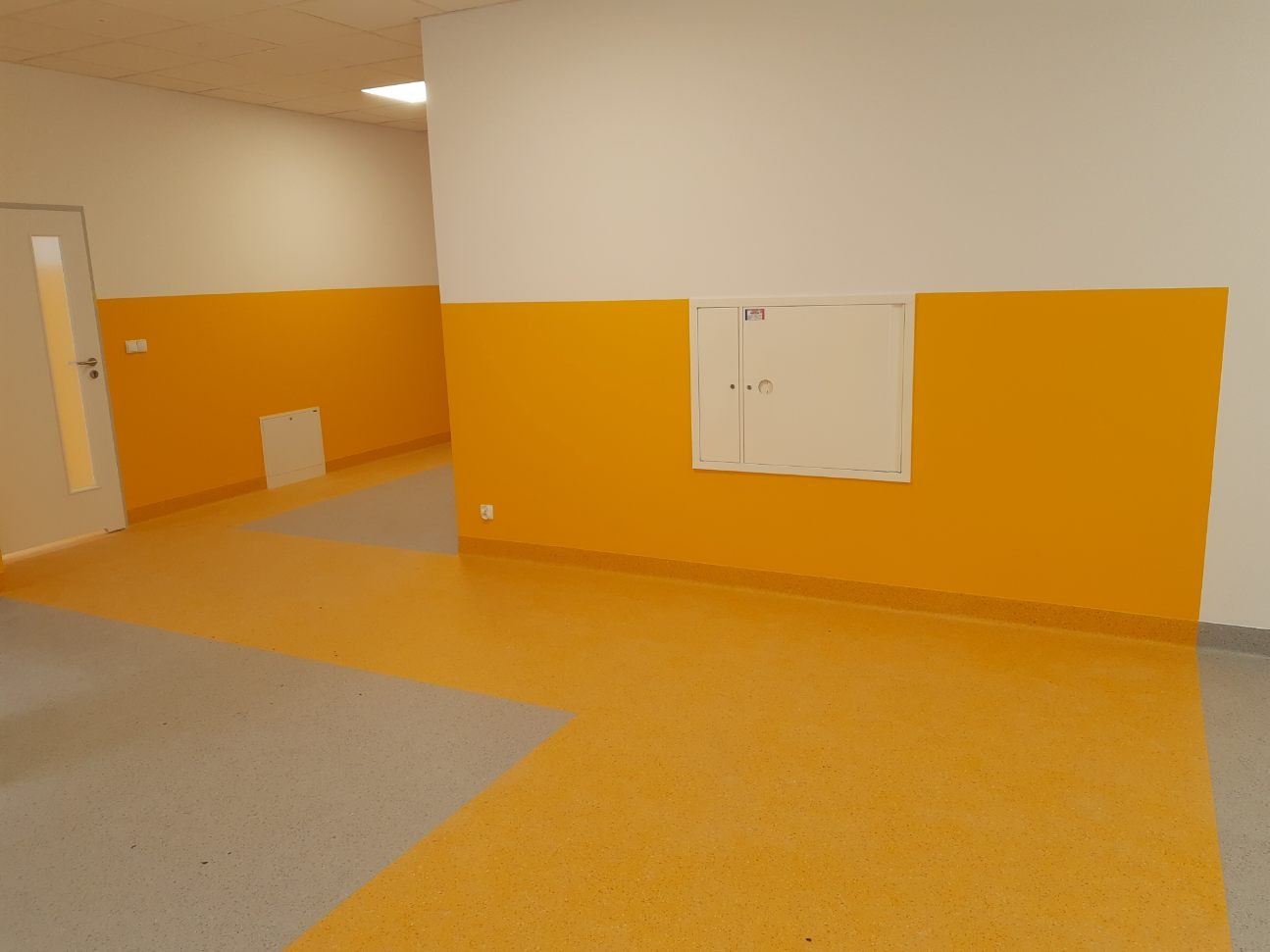 Nowa, dwukolorowa, żółto-szara podłoga z wykładziny w korytarzu biurowym, z żółtymi ścianami do połowy wysokości i białymi powyżej. Widoczna biała szafka rewizyjna na ścianie.