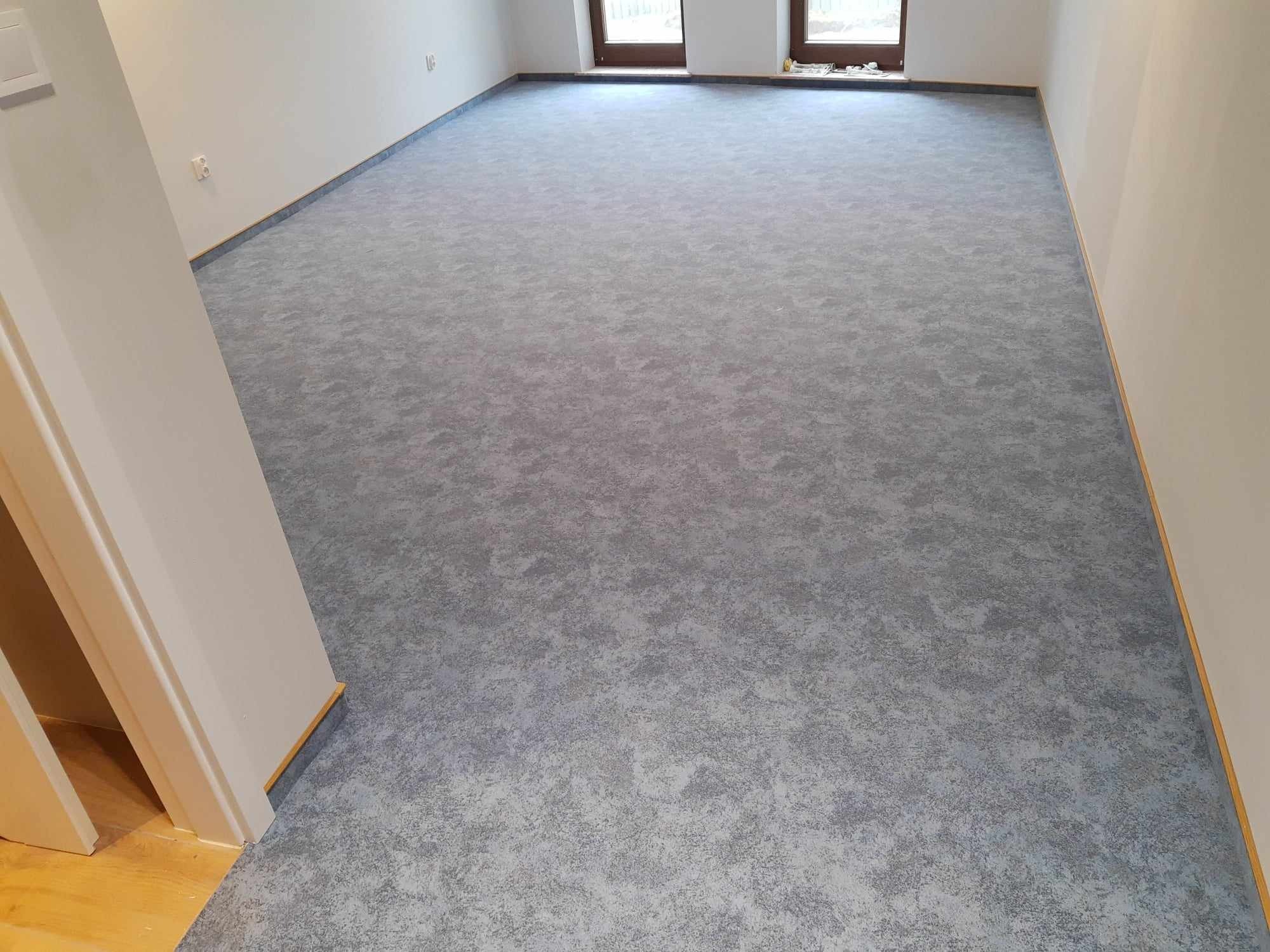 Pokój z nową, szarą wykładziną dywanową o wzorze melanżowym, widoczne listwy przypodłogowe i fragment drewnianej podłogi przy drzwiach.