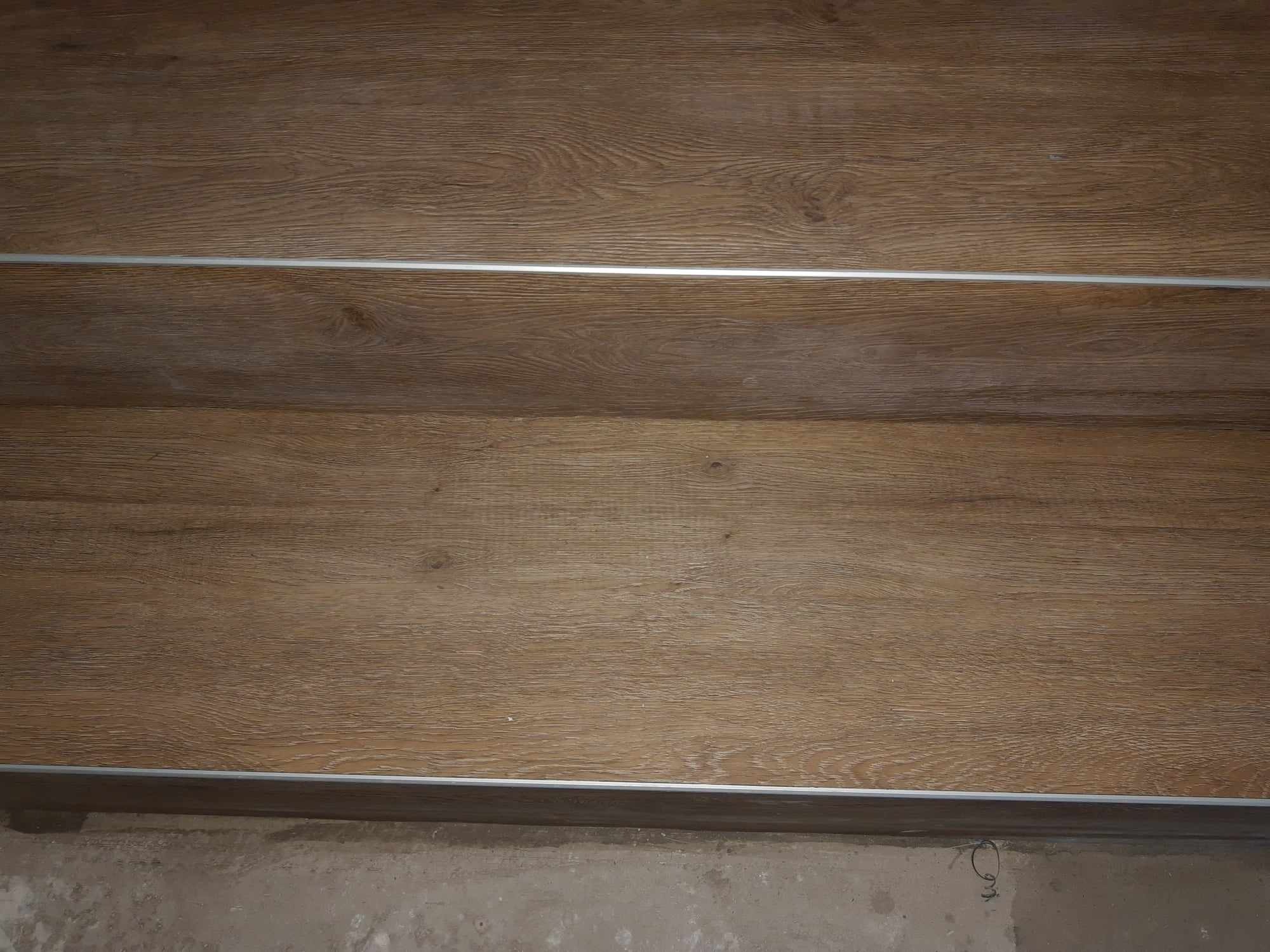 Fragment schodów wykończonych panelami imitującymi jasne drewno, z aluminiowymi listwami zabezpieczającymi krawędzie, widoczny surowy beton u podstawy.