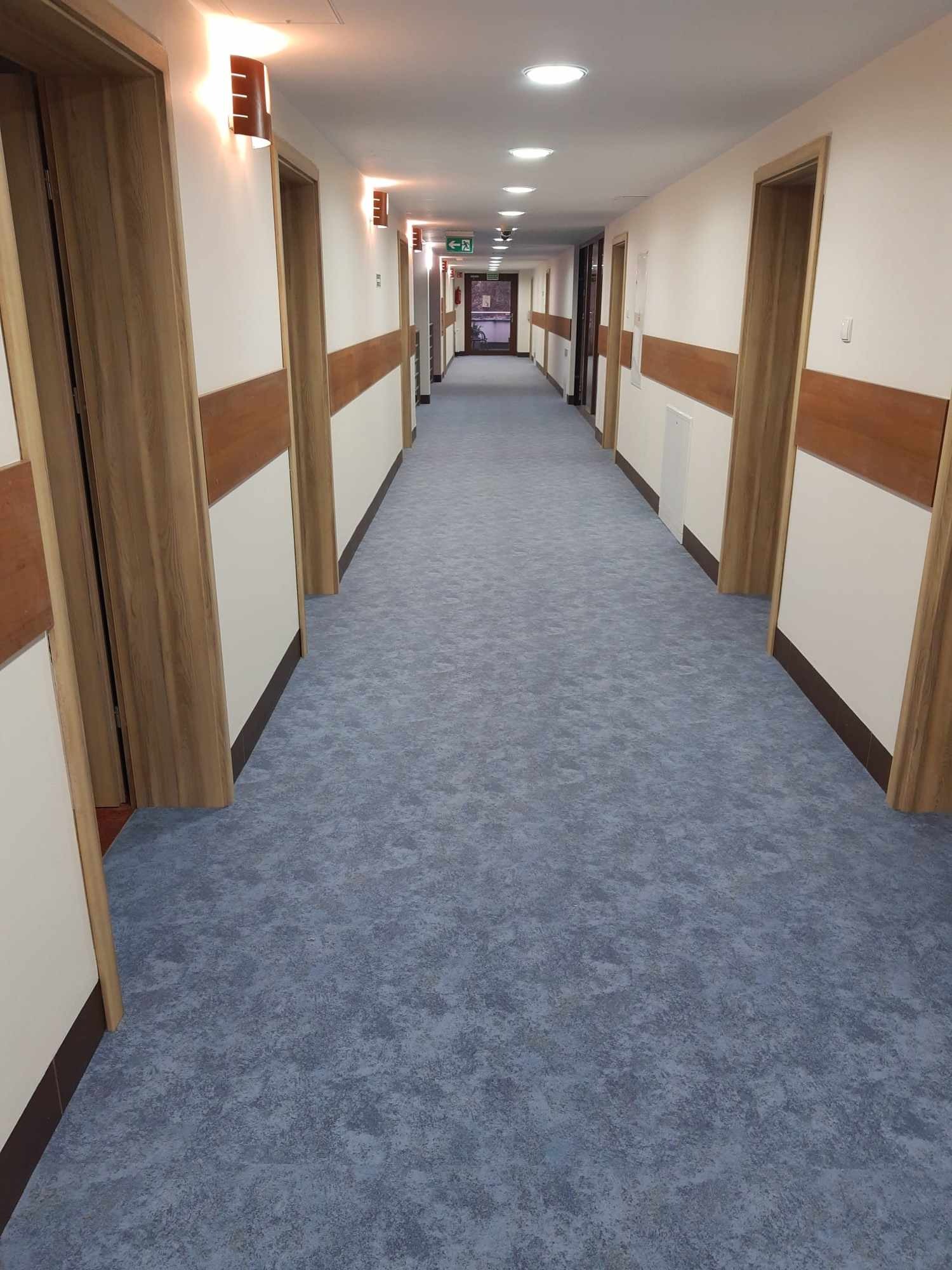 Długi korytarz w budynku użyteczności publicznej z szarą wykładziną dywanową o wzorzystej strukturze, drewnianymi drzwiami i oświetleniem sufitowym.