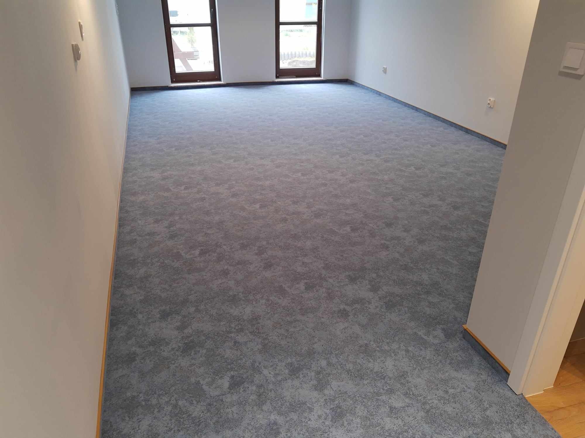 Pokój z nową, szarą wykładziną dywanową o nieregularnym wzorze, widoczne dwa okna z brązowymi ramami i białe ściany z listwami przypodłogowymi.
