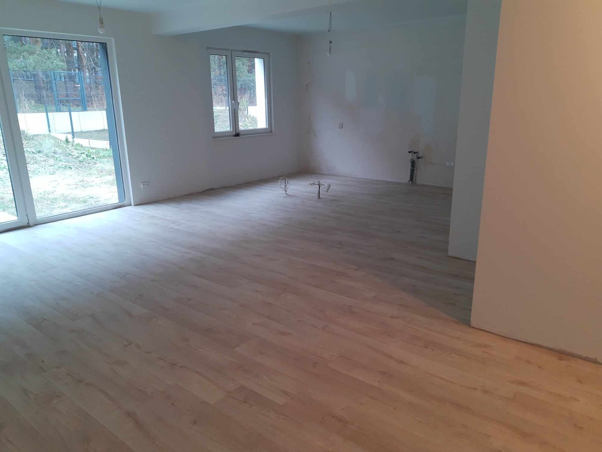 Jasne panele podłogowe ułożone w nowym, pustym pomieszczeniu z dużymi oknami balkonowymi i niedokończonymi ścianami.