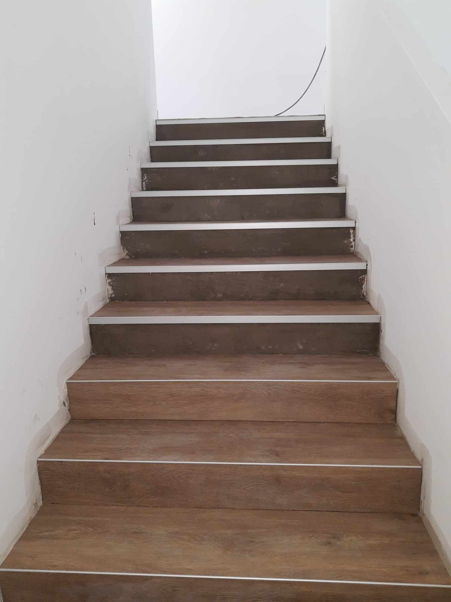 Schody wyłożone panelami imitującymi drewno, z metalowymi listwami na krawędziach, widoczne ślady prac wykończeniowych na ścianach.
