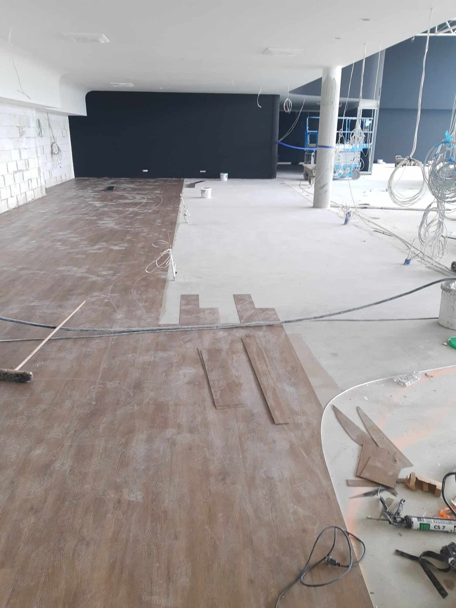 Połowa pomieszczenia z brązowymi panelami podłogowymi, druga połowa z surową betonową podłogą, widoczne narzędzia i materiały budowlane, w tle czarna ściana i instalacje elektryczne