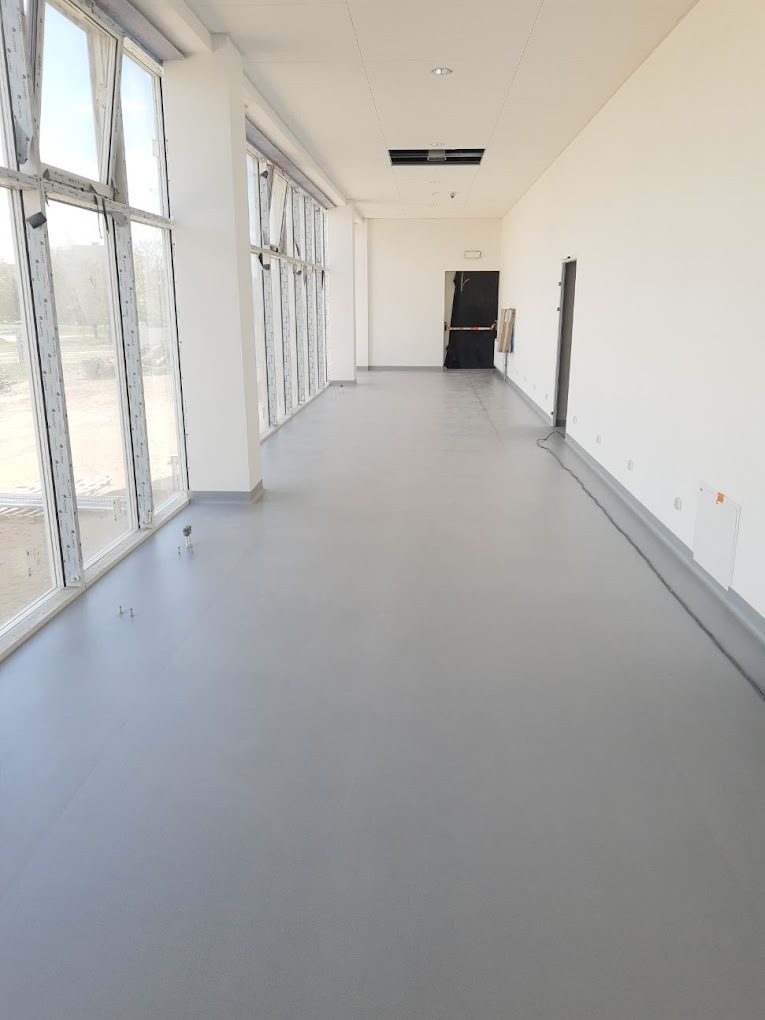Długi korytarz z szarą, jednolitą posadzką, duże okna po lewej stronie wpuszczające naturalne światło, widoczne drzwi na końcu korytarza.